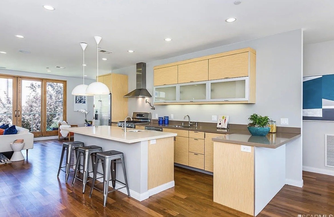 Modern kitchen with light wood cabinets, white countertops, stainless steel appliances, and a breakfast bar with four gray metal bar stools. There is a sliding glass door leading to a balcony with snow outside.