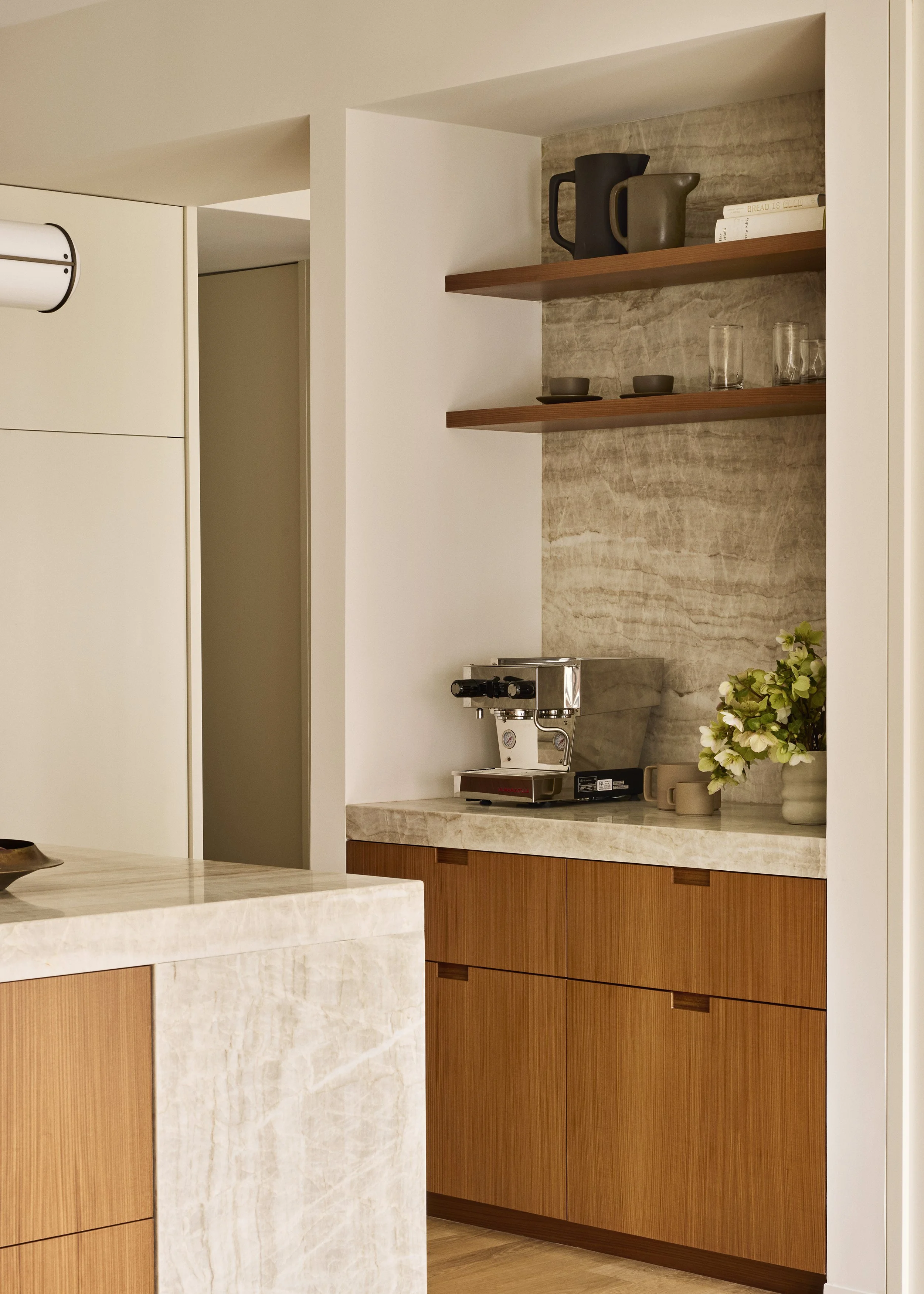 A modern kitchen corner with floating wooden shelves holding cups, glasses, and a book, a coffee machine on a marble countertop, and a vase with white flowers.