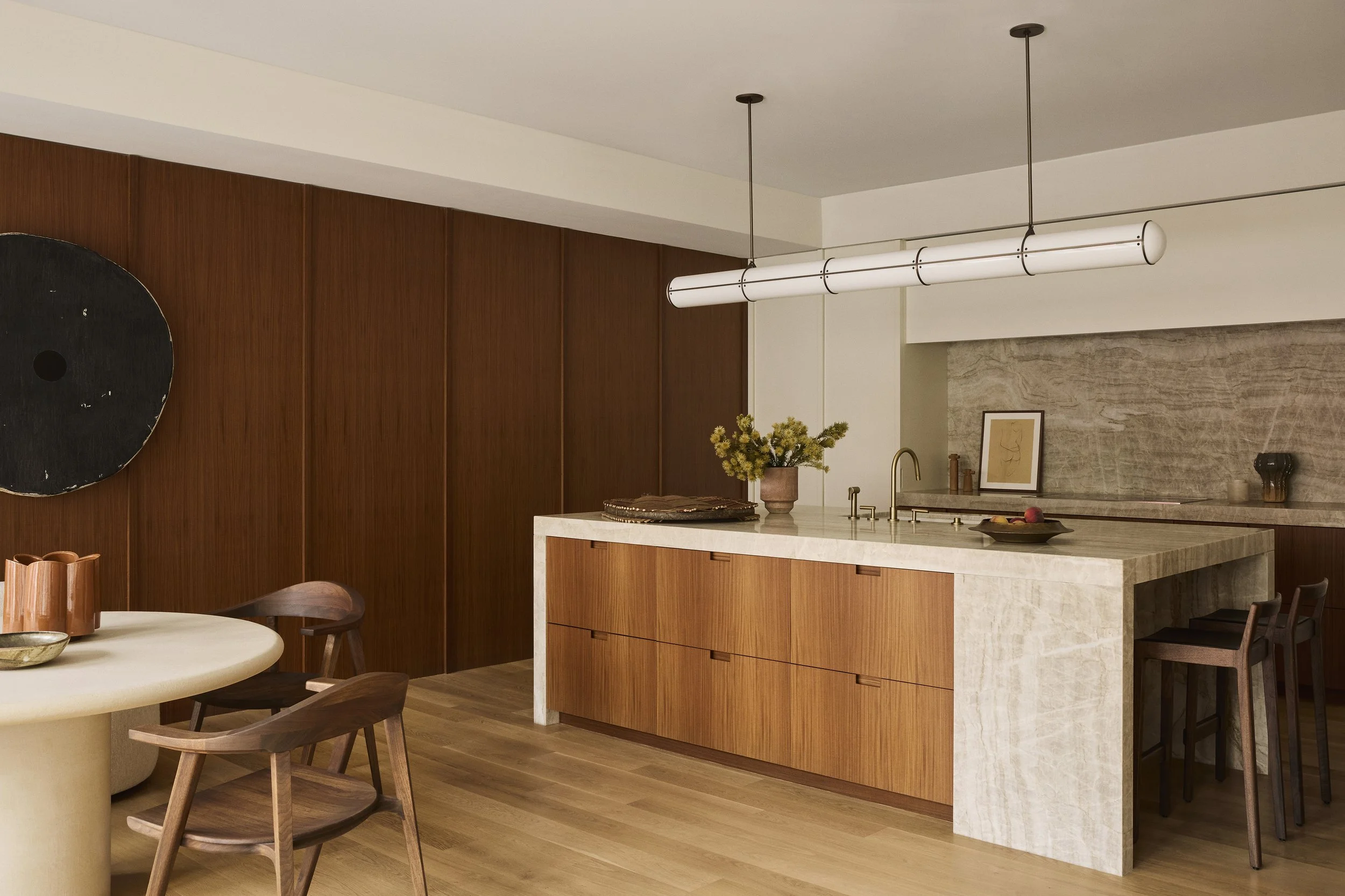Modern kitchen with wood-paneled wall, marble island, and minimalist decor, including a round dining table with wooden chairs and a large black circular wall art.