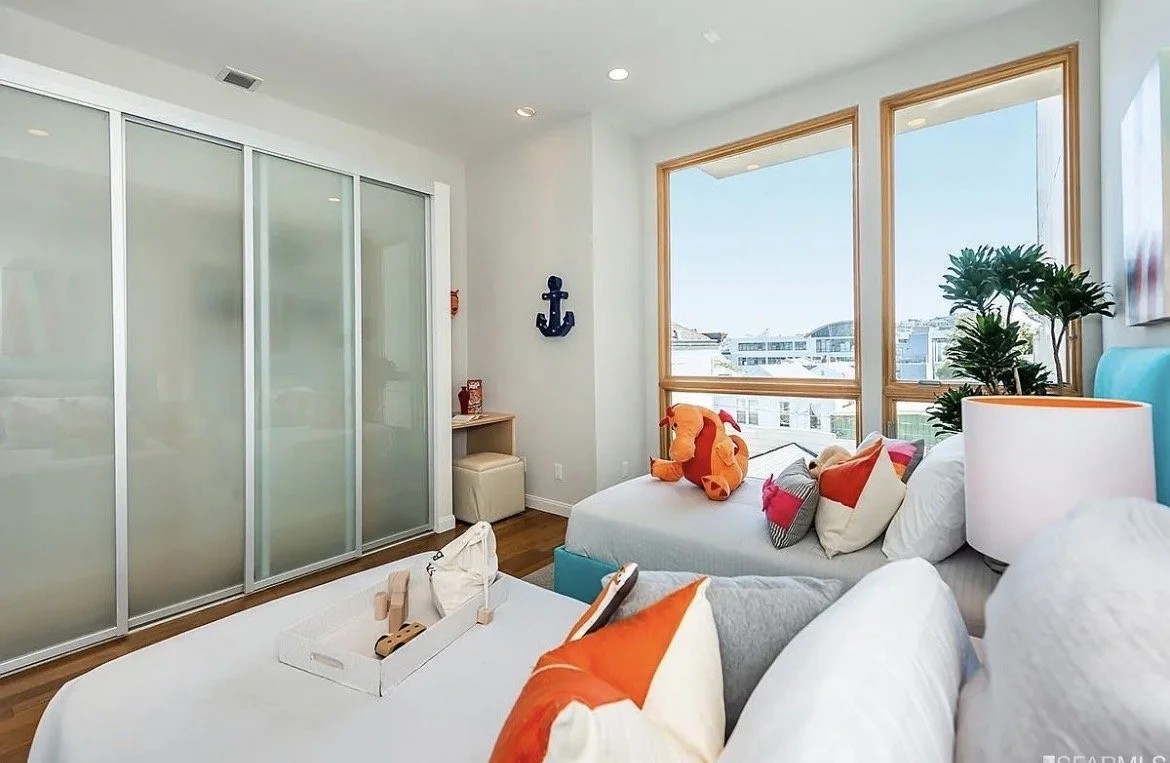 Bright bedroom with large windows showing city view, decorated with colorful pillows, plush toy, a lamp, potted plants, and a large closet with sliding frosted glass doors.