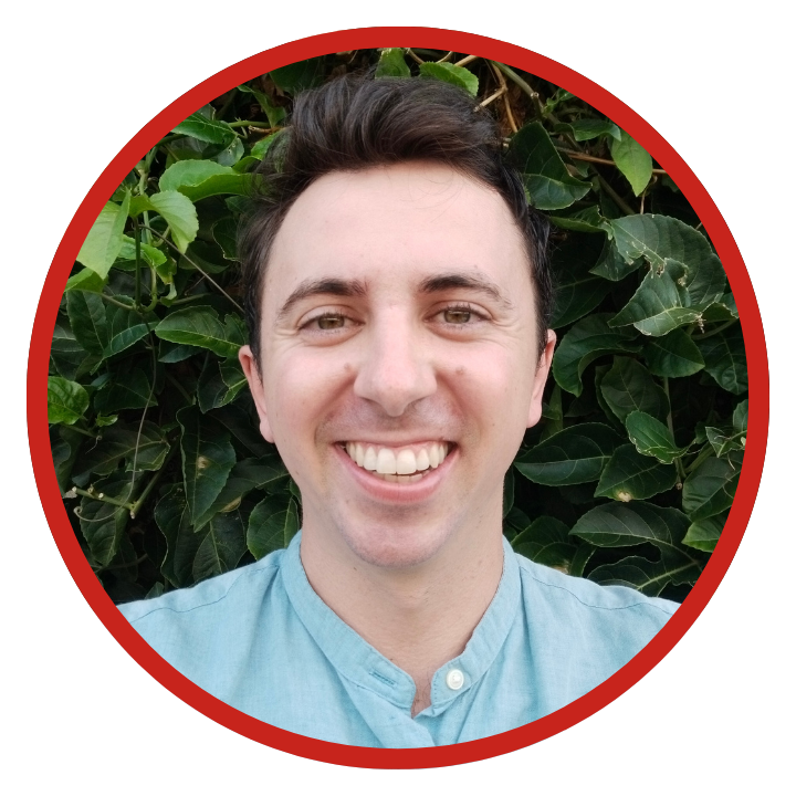A young man with dark hair smiling in front of green leafy plants, wearing a light blue shirt.