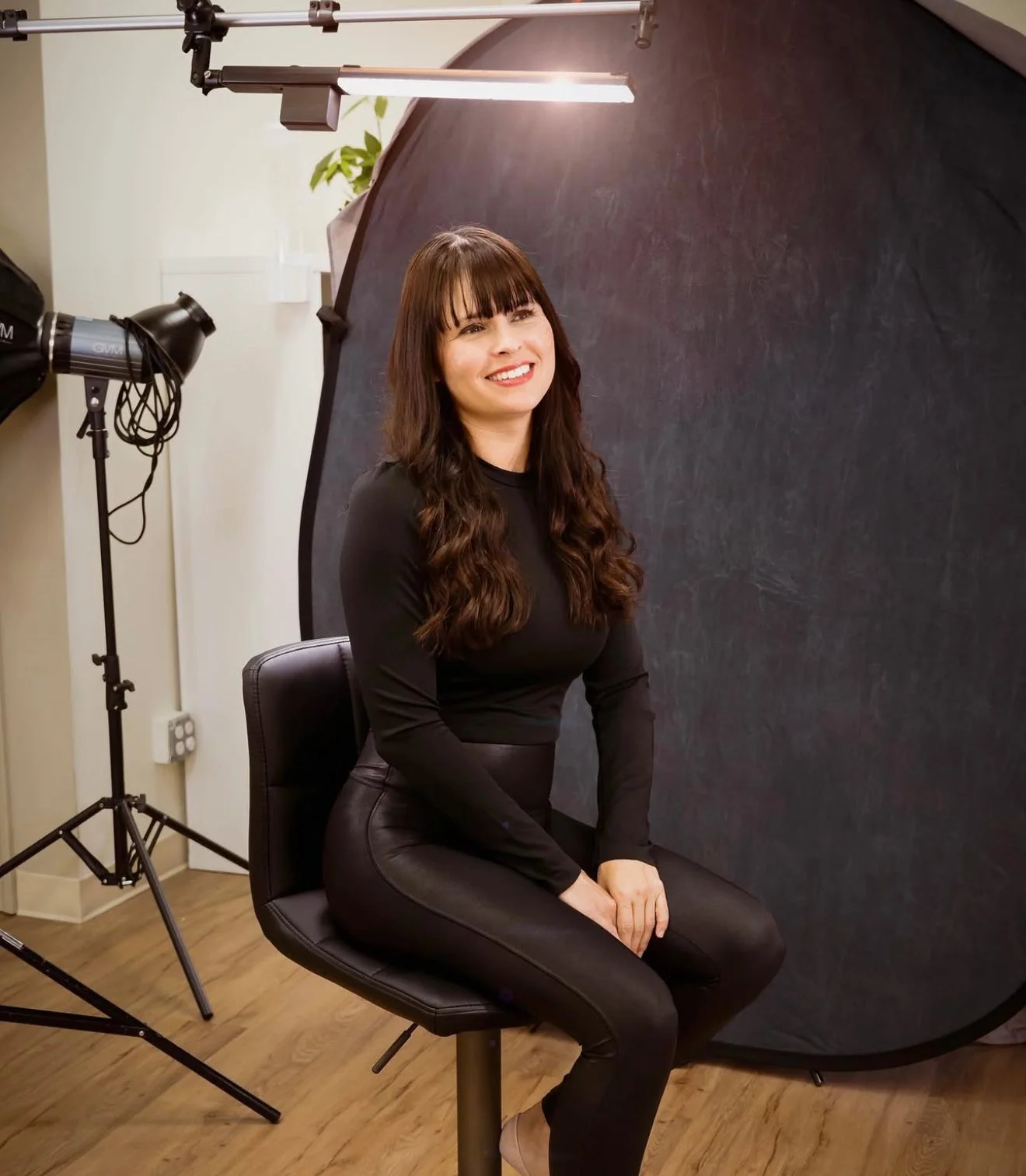 Woman with long wavy brown hair in black outfit seated in a photography studio with a dark backdrop and professional lighting.