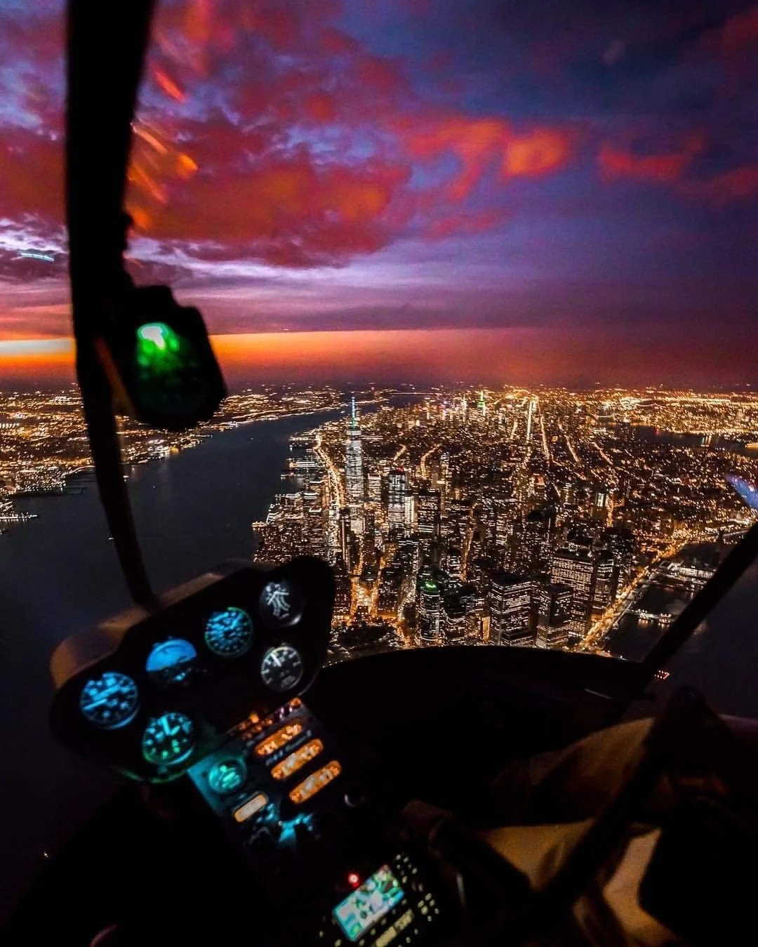 View from a helicopter over a city at sunset with a colorful sky, illuminated buildings, and streets, with helicopter controls and instrument panel in the foreground.