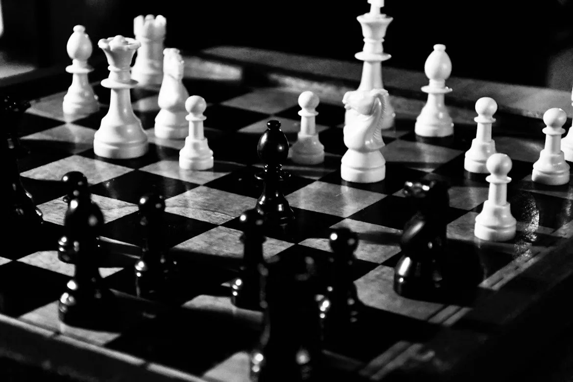 A black and white photograph of a chessboard with white and black chess pieces in mid-game.
