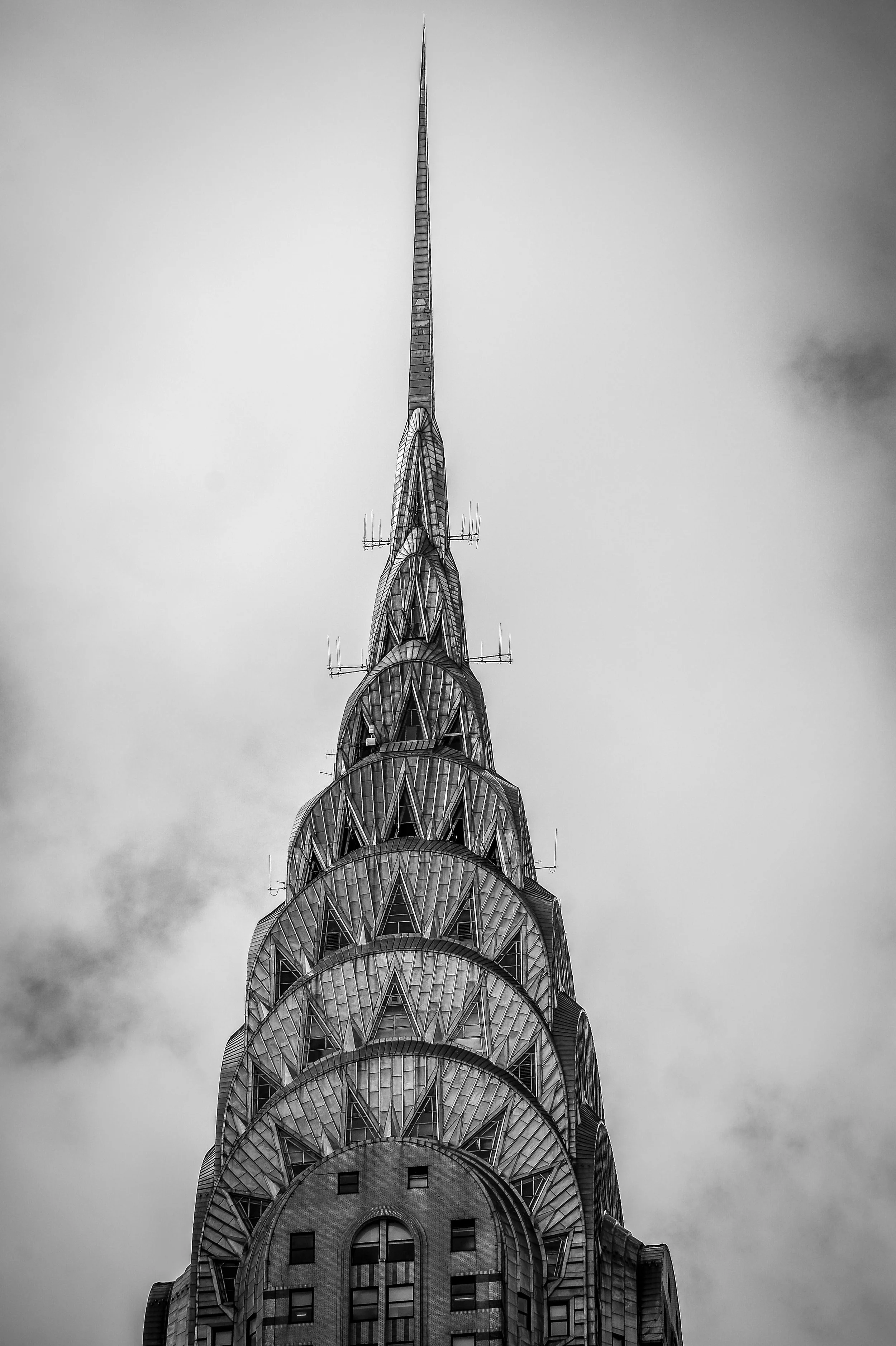 Black and white photo of the top section of the Chrysler Building in New York City, featuring its spire and distinctive Art Deco design.