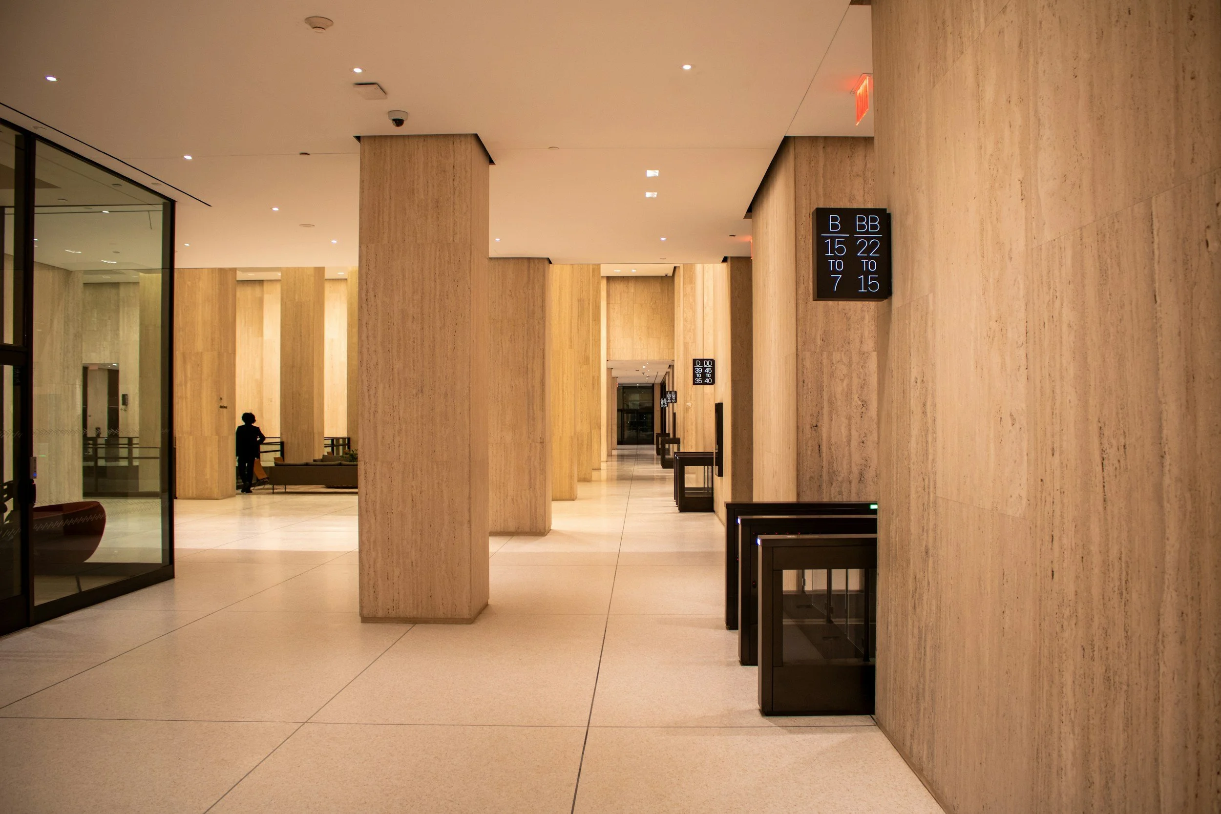 Modern airport terminal with beige stone walls and columns, digital gate information screens, and a person waiting in the distance.