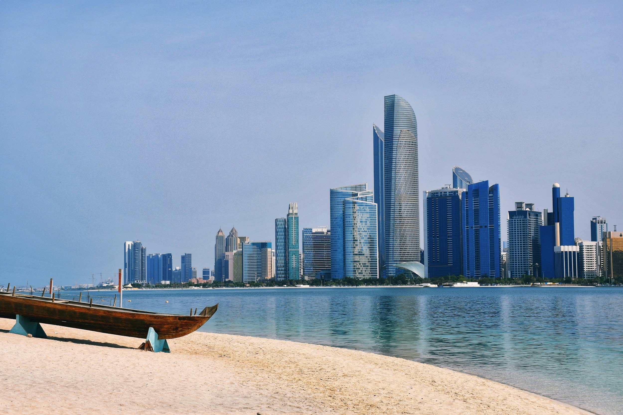 View of a modern city skyline with tall skyscrapers along the waterfront, seen from a sandy beach with a small wooden boat in the foreground.