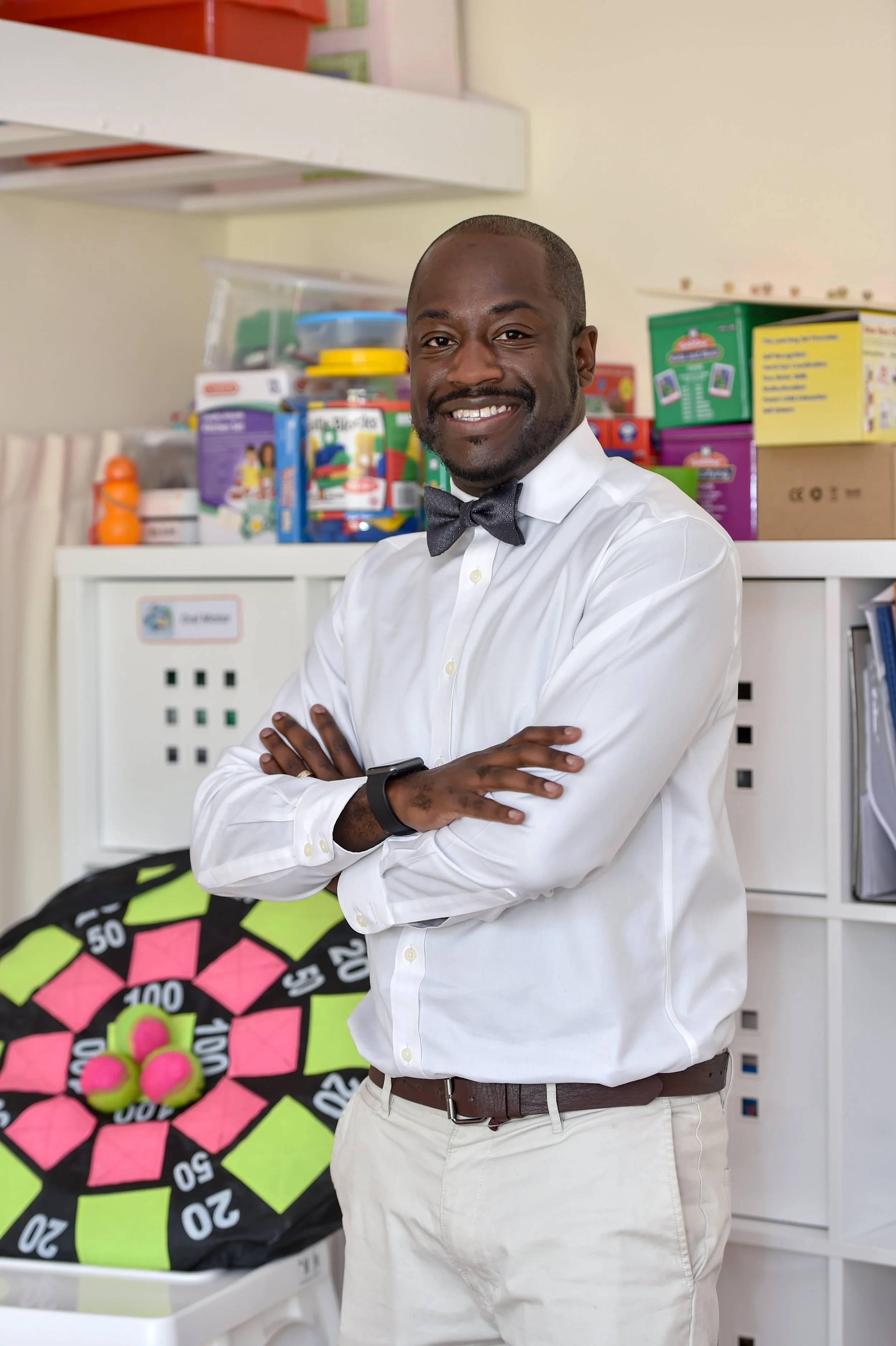 A man dressed in a white shirt and black bow tie, standing with his arms crossed and smiling in an office or classroom with colorful toys and games on shelves behind him, including a pink and green tennis ball dartboard on a desk.