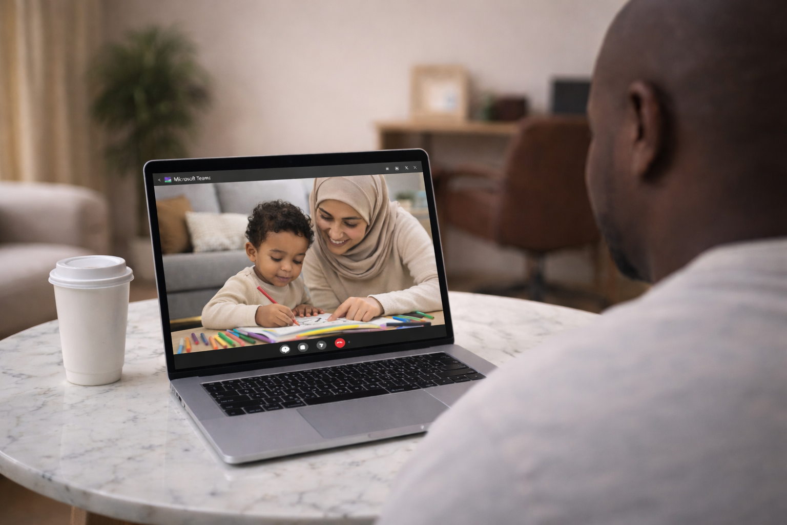 A man on a video call with a woman and a child on a laptop, sitting at a marble table with a coffee cup nearby.