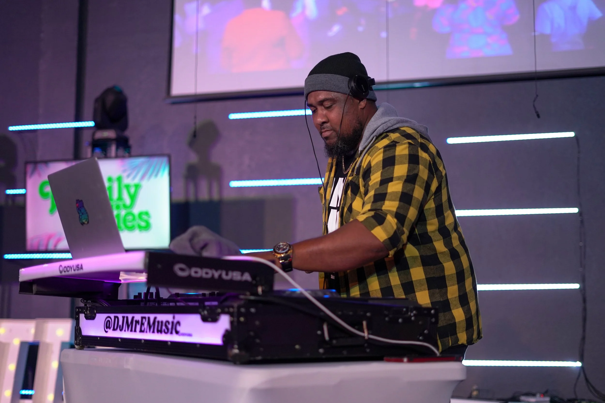 A DJ wearing a black beanie, gray hoodie, and yellow plaid shirt, mixes music with DJ equipment in a dimly lit room with neon lights and a microphone stand.