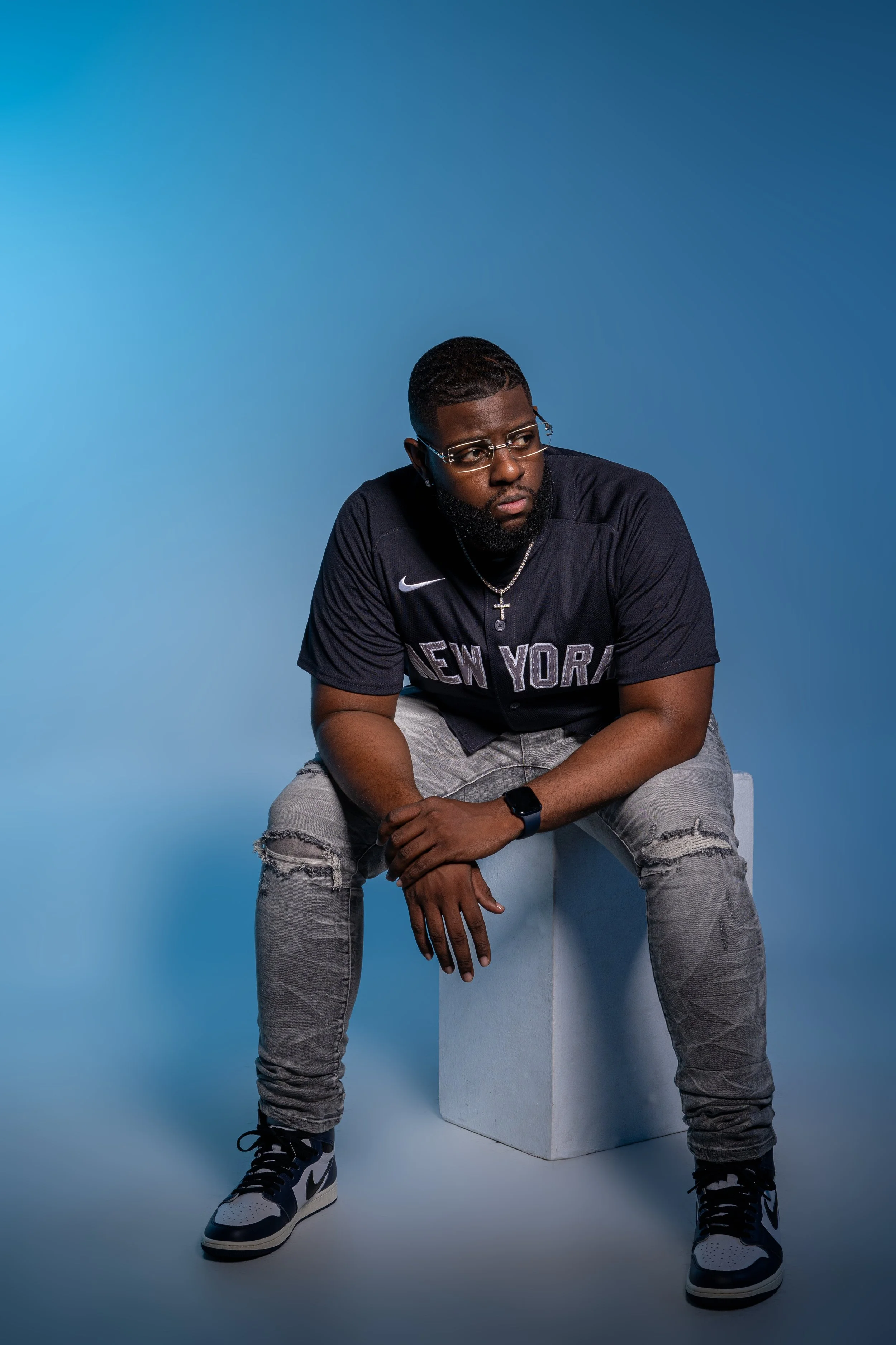 A man wearing glasses, a black New York sports jersey, grey ripped jeans, and Nike sneakers, sitting on a white cube against a blue background.