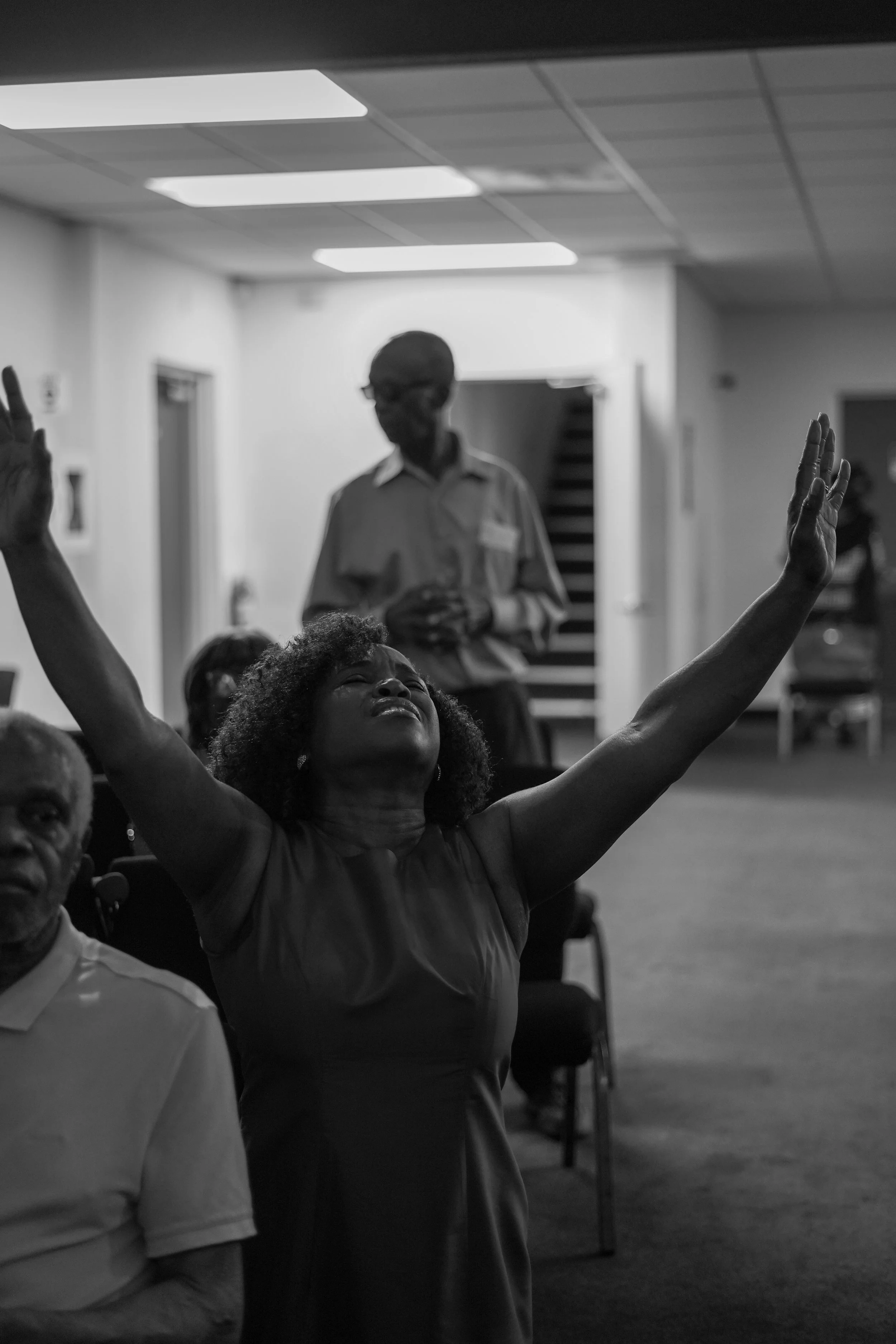 A woman with curly hair has her eyes closed and arms raised in front of a group, with a man in the background standing with hands clasped in an indoor setting.