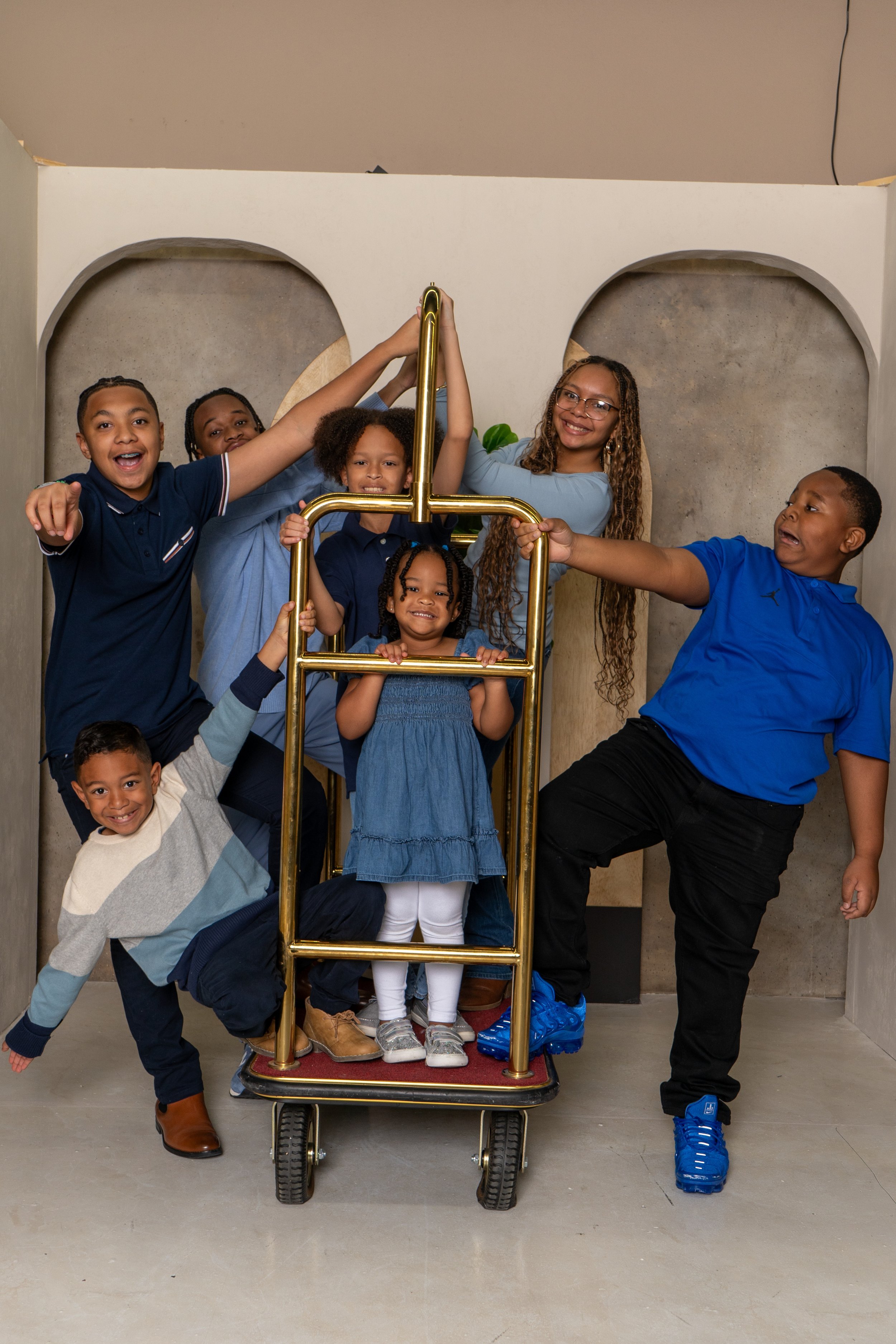 Group of laughing children and two adults posing on and around a gold-colored cart inside a room with a plain background.