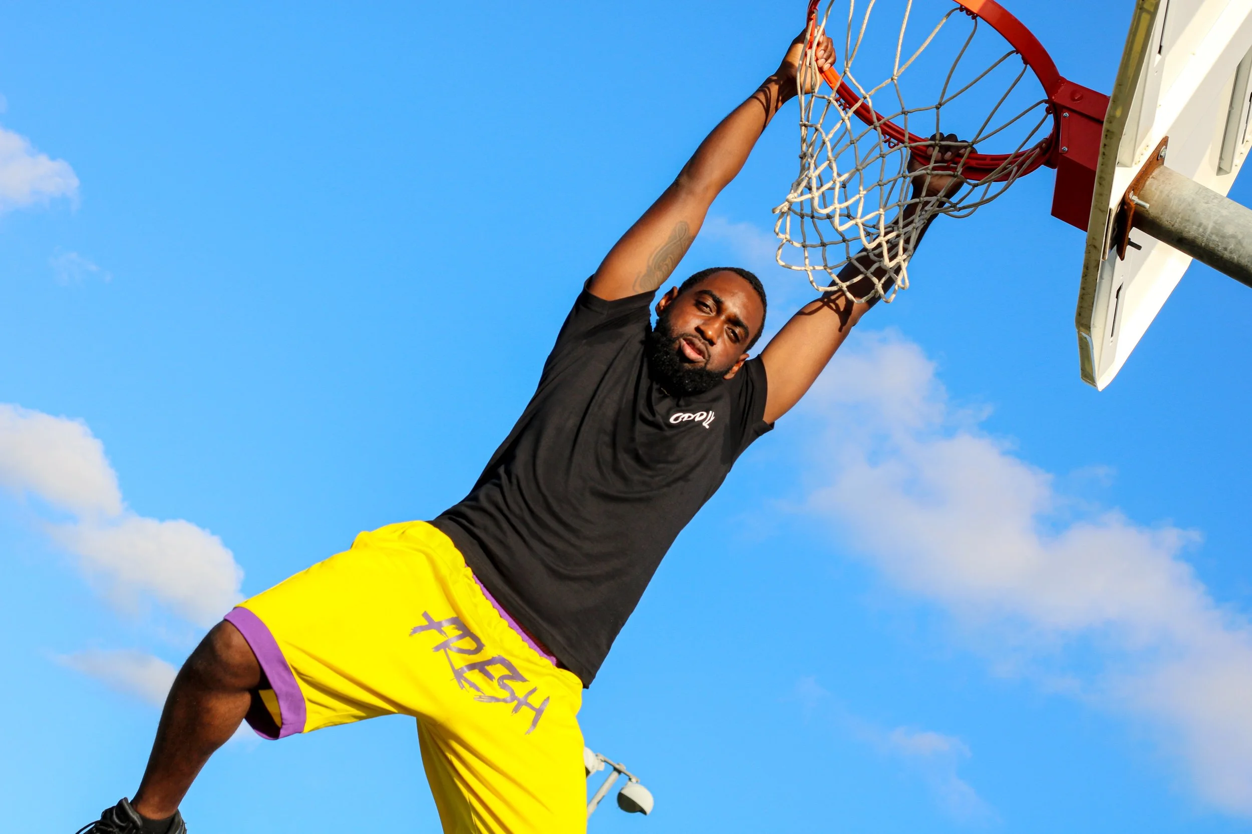 A man in yellow shorts and a black T-shirt is hanging from a basketball hoop, about to dunk a basketball.