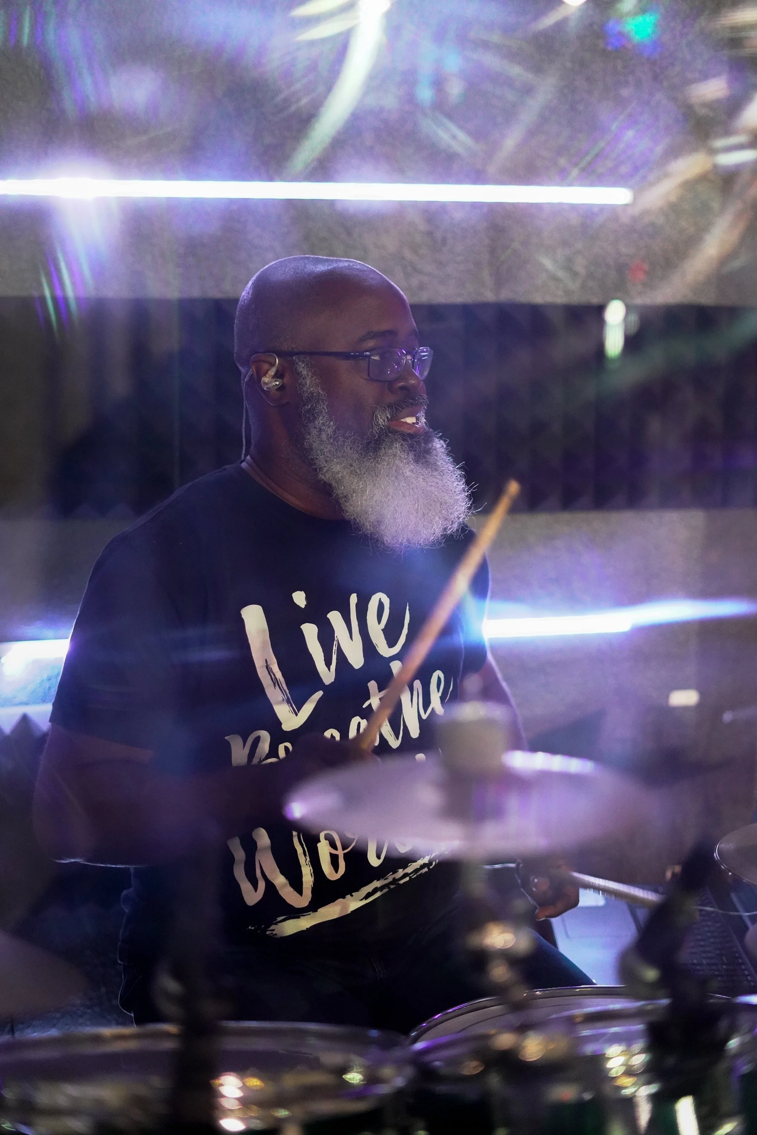 A bearded man with glasses and an in-ear monitor playing drums in a dimly lit setting with colorful lights.