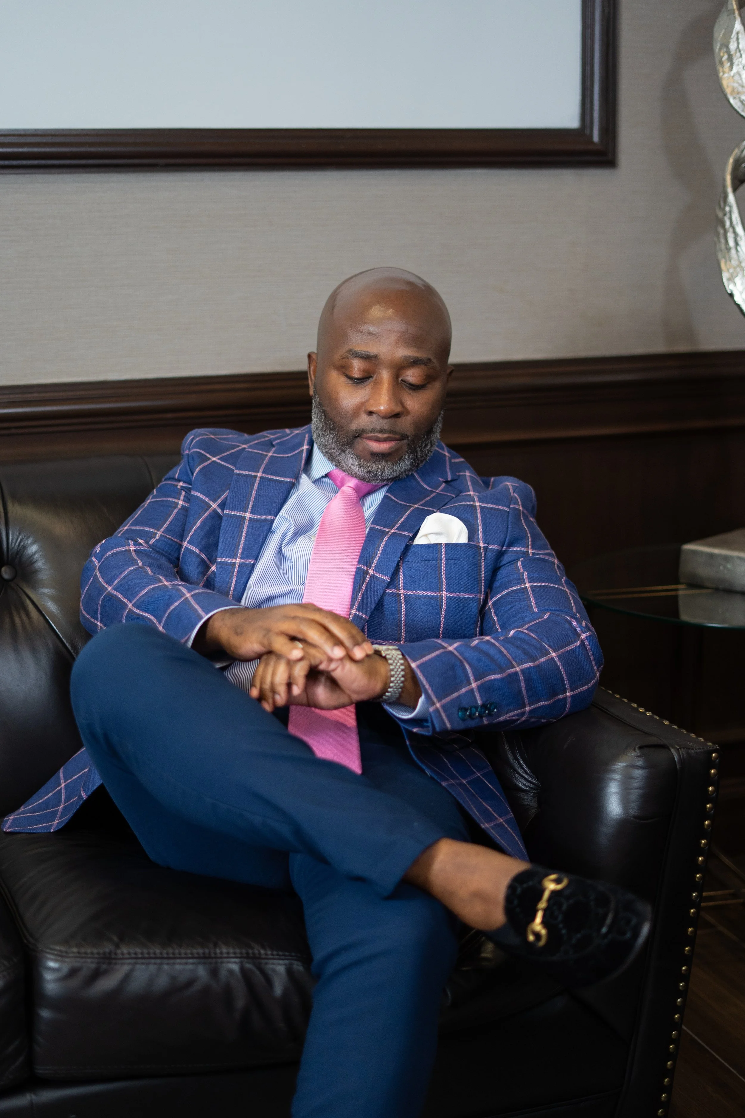 A man sitting on a black leather sofa, dressed in a blue checkered suit with pink tie, white shirt, and black shoes, looking at his watch, in a modern, elegant room.
