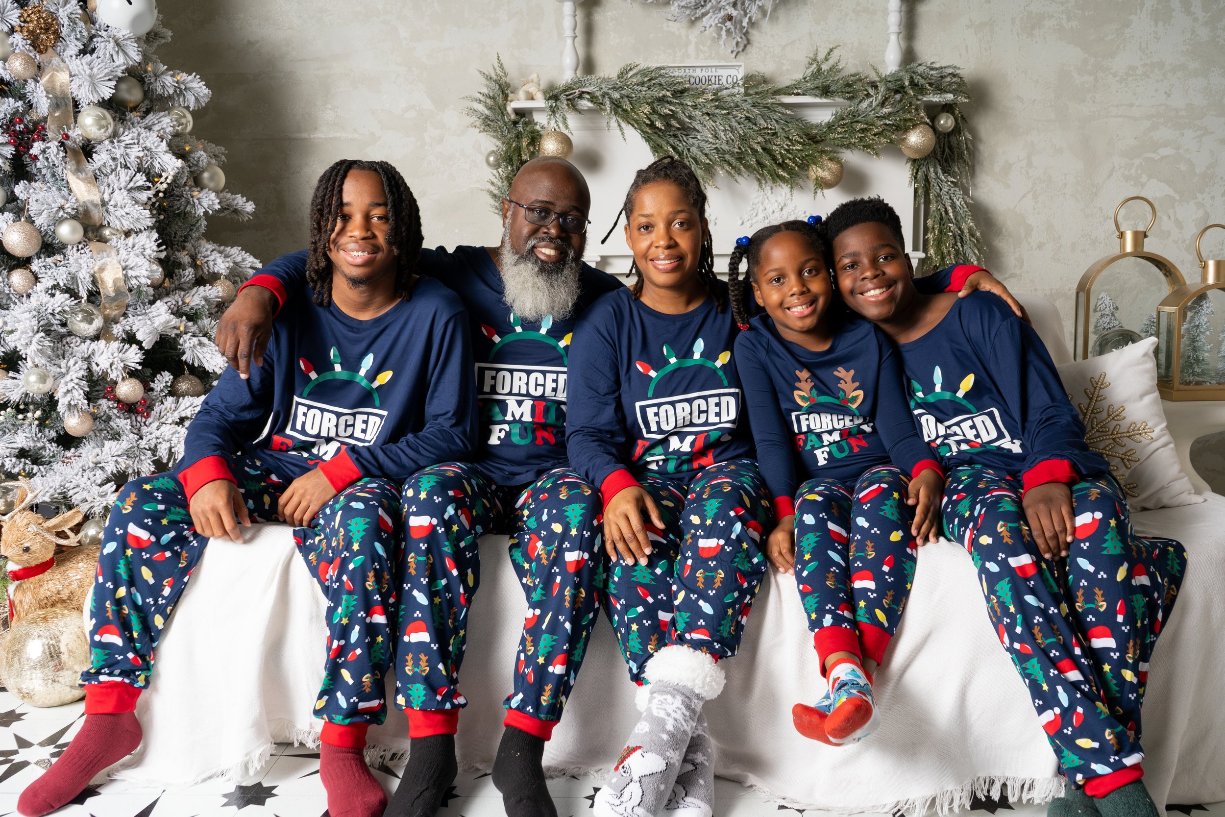 A family of six sitting on a white couch in front of a decorated Christmas tree, all wearing matching Christmas pajamas with festive designs, smiling and celebrating during Christmas.