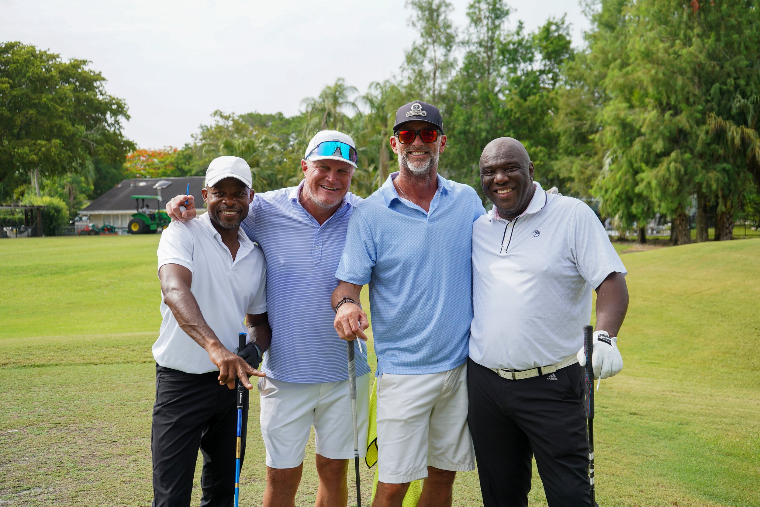 Four men smiling and posing on a golf course, holding golf clubs, with green trees and a golf cart in the background.