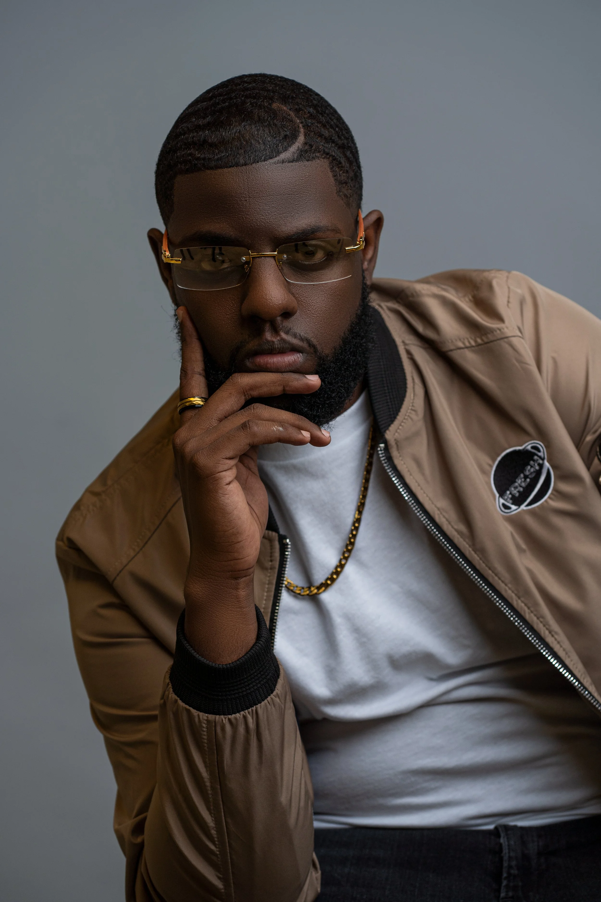 Portrait of a young man with dark skin, wearing glasses, a beige jacket, a white t-shirt, a gold necklace, and rings, posing with his hand on his chin against a gray background.