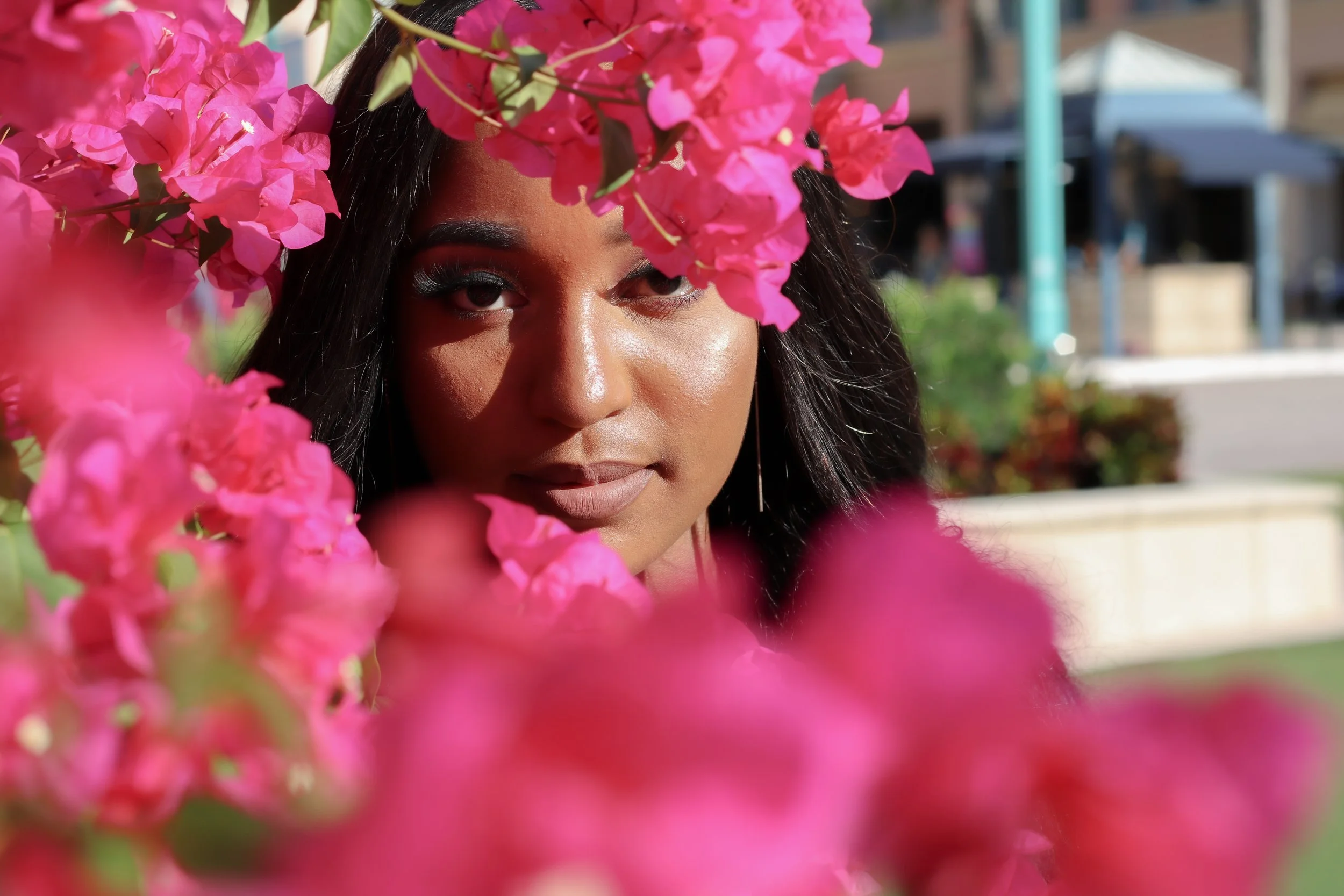 A woman with dark hair and makeup, partly obscured by pink bougainvillea flowers, outdoors with a blurred background of a park or plaza.