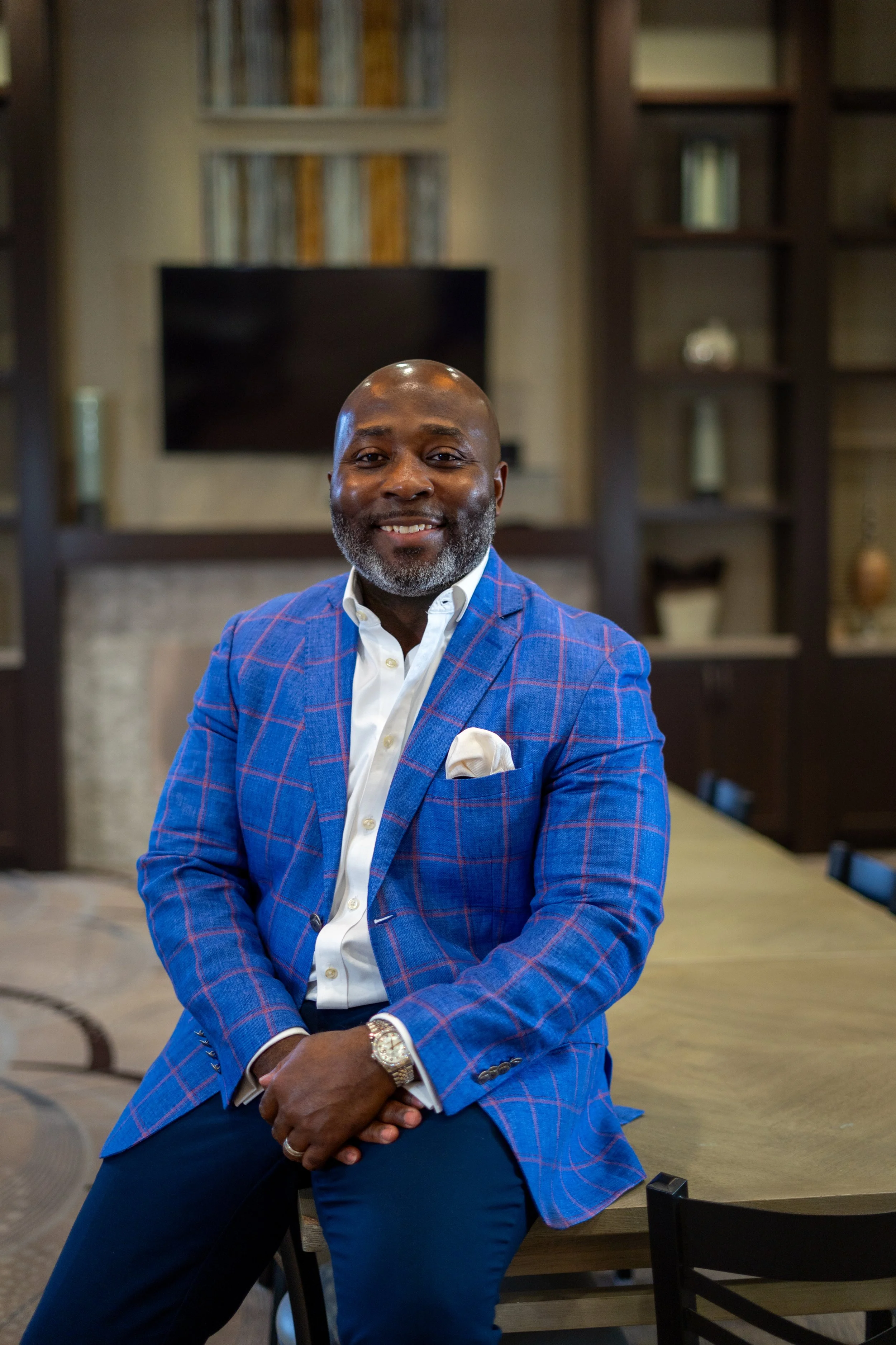 A man in a blue checkered blazer and white shirt, sitting on a table, smiling in a room with bookshelves and a television behind him.