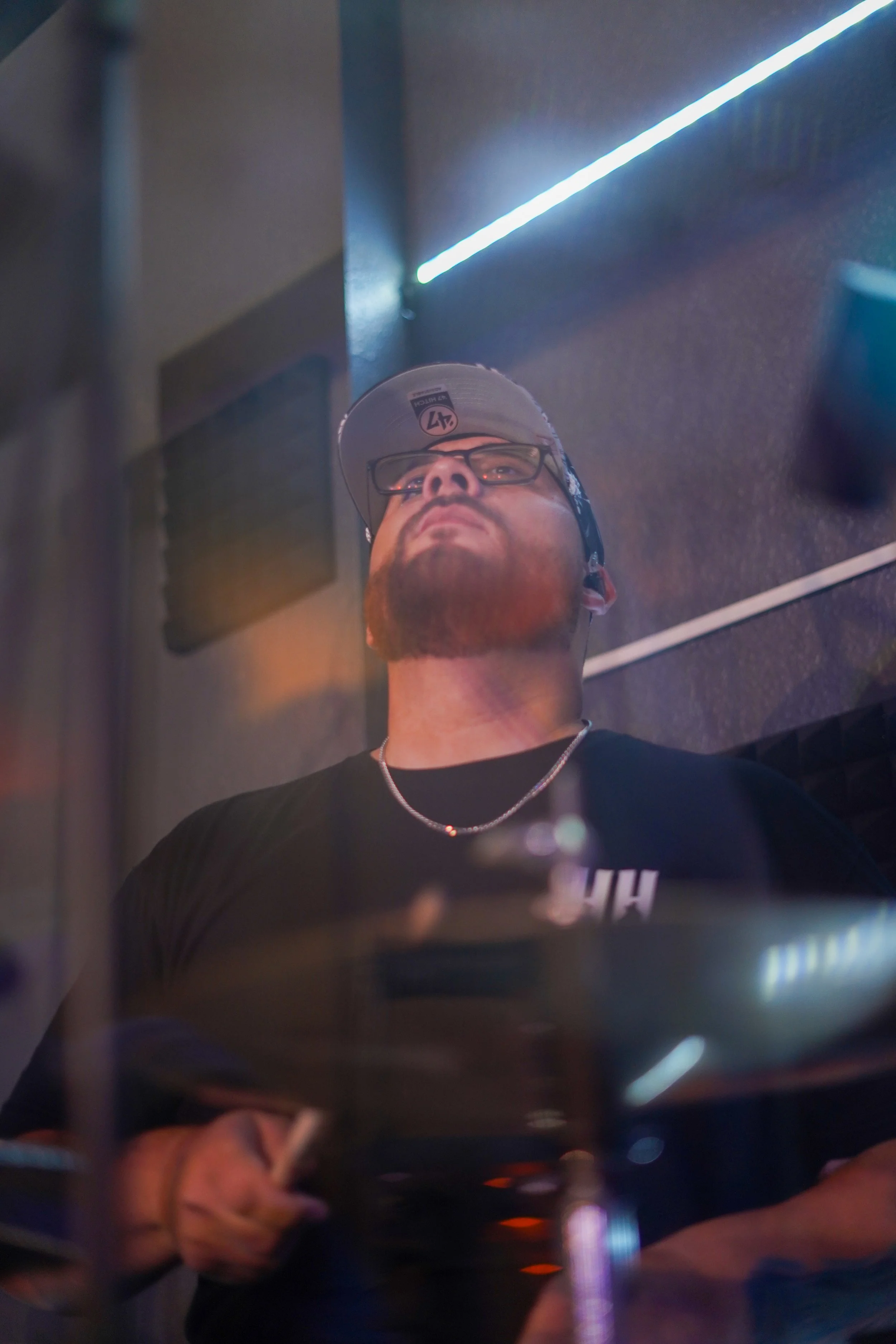 A person with glasses, a beard, and a baseball cap is playing drums with a focused expression, with blue and warm lighting in a music studio