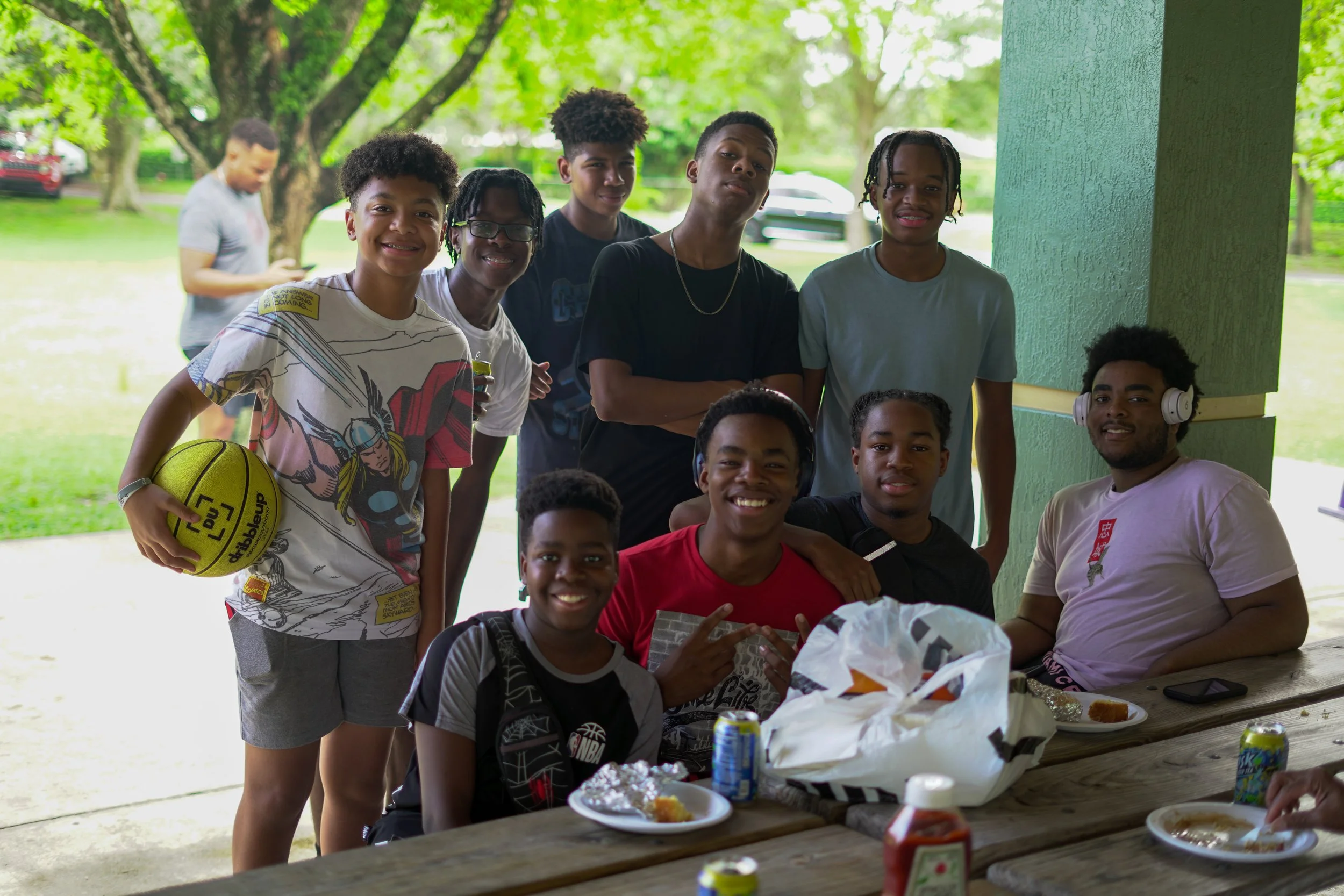 A group of eleven young people, mostly teenagers, gathered around a picnic table outdoors under a pavilion. Some are sitting, smiling, and making hand signs, while others are standing behind. The group includes boys and girls with diverse hairstyles and clothing. The table has plates of food, drinks, and a large white bag. Two people can be seen in the background near trees, one focusing on their phone. The scene appears to be a casual outdoor gathering or picnic on a sunny day.