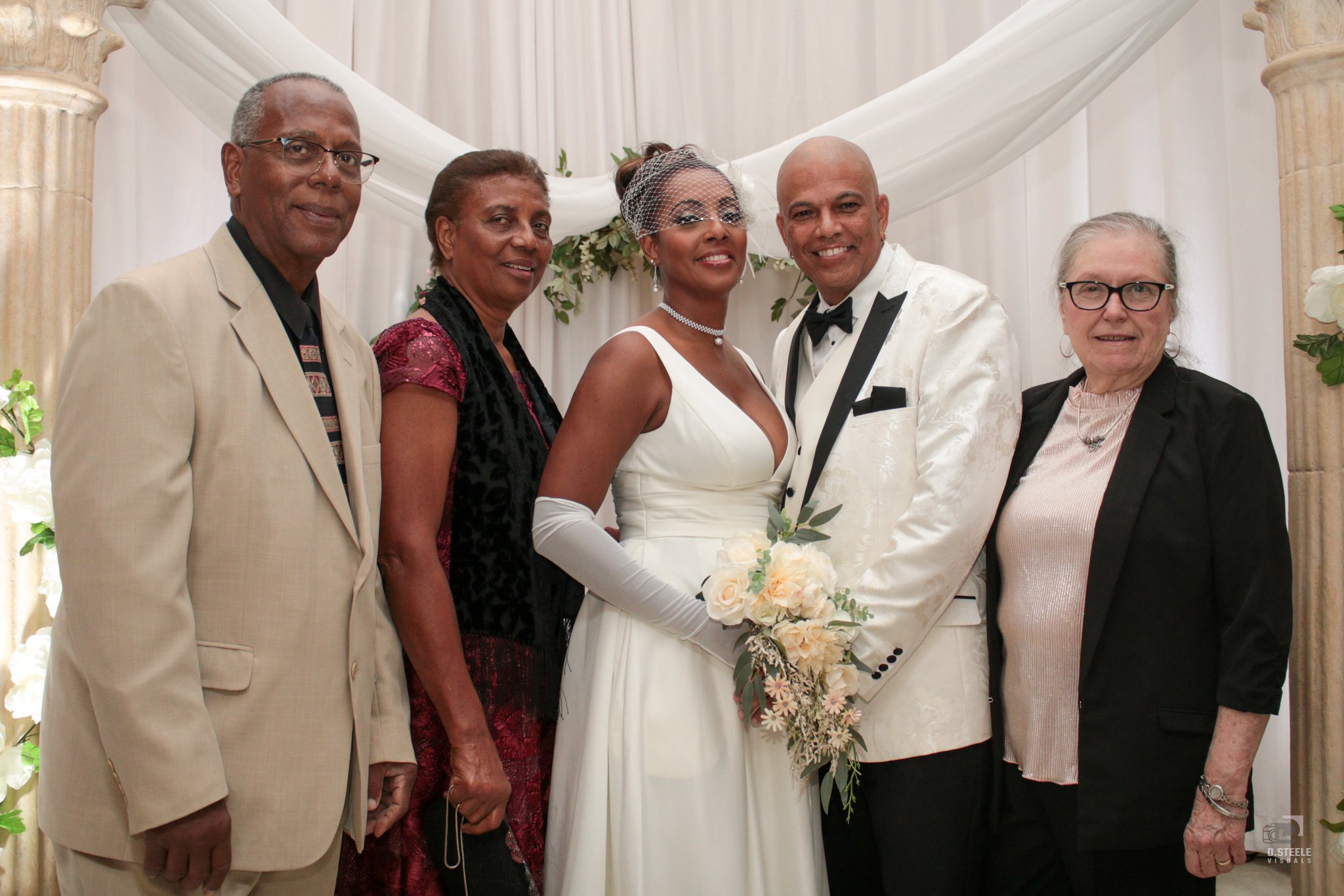 A wedding celebration with five people standing together, including a bride in a white wedding gown holding a bouquet, and a groom in a white tuxedo. Four others, two men and two women, are standing beside them, dressed in formal attire, in front of a decorative backdrop with white fabric and greenery.