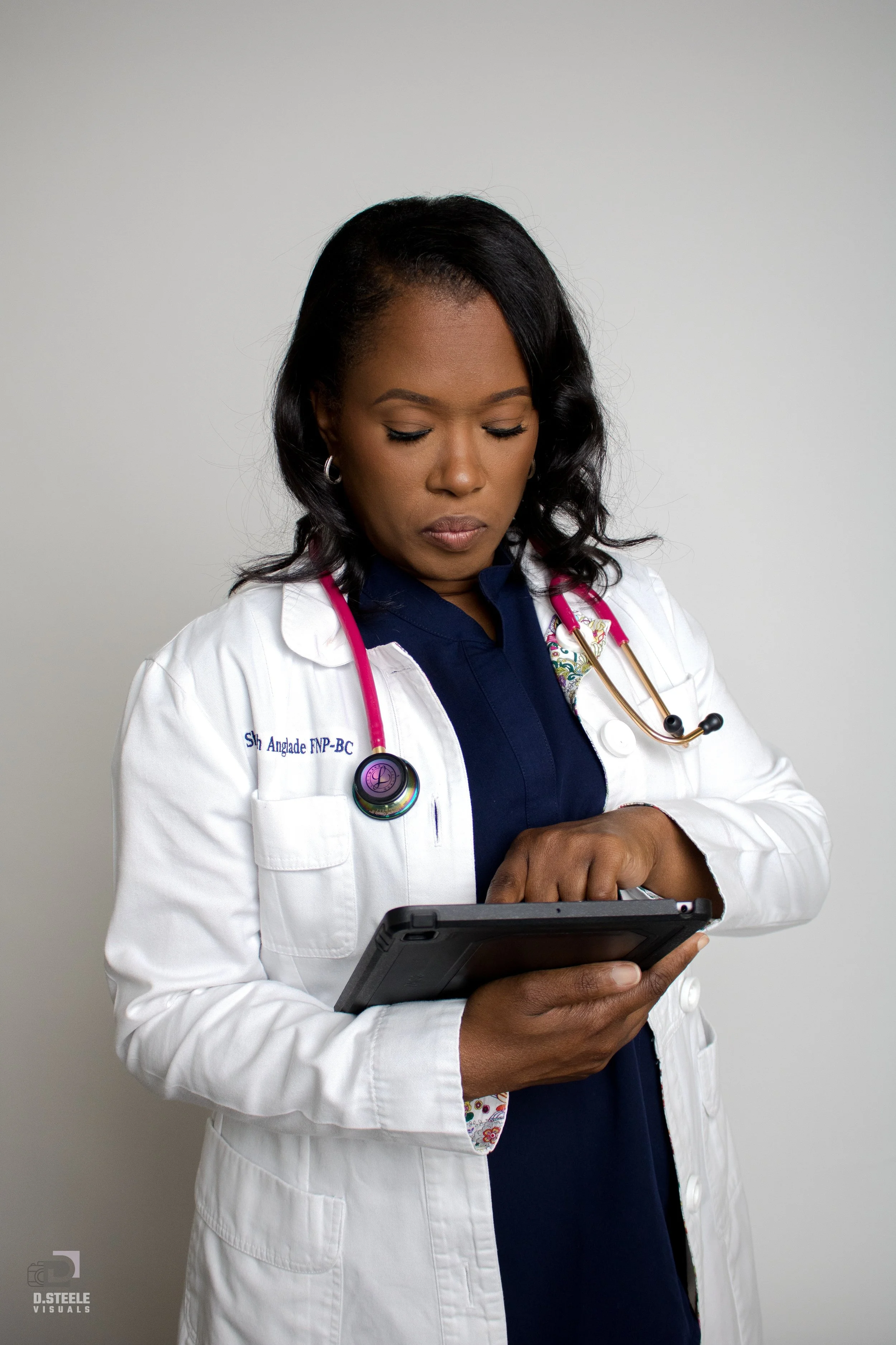 Medical professional wearing a white lab coat and stethoscope using a tablet device against a plain background.