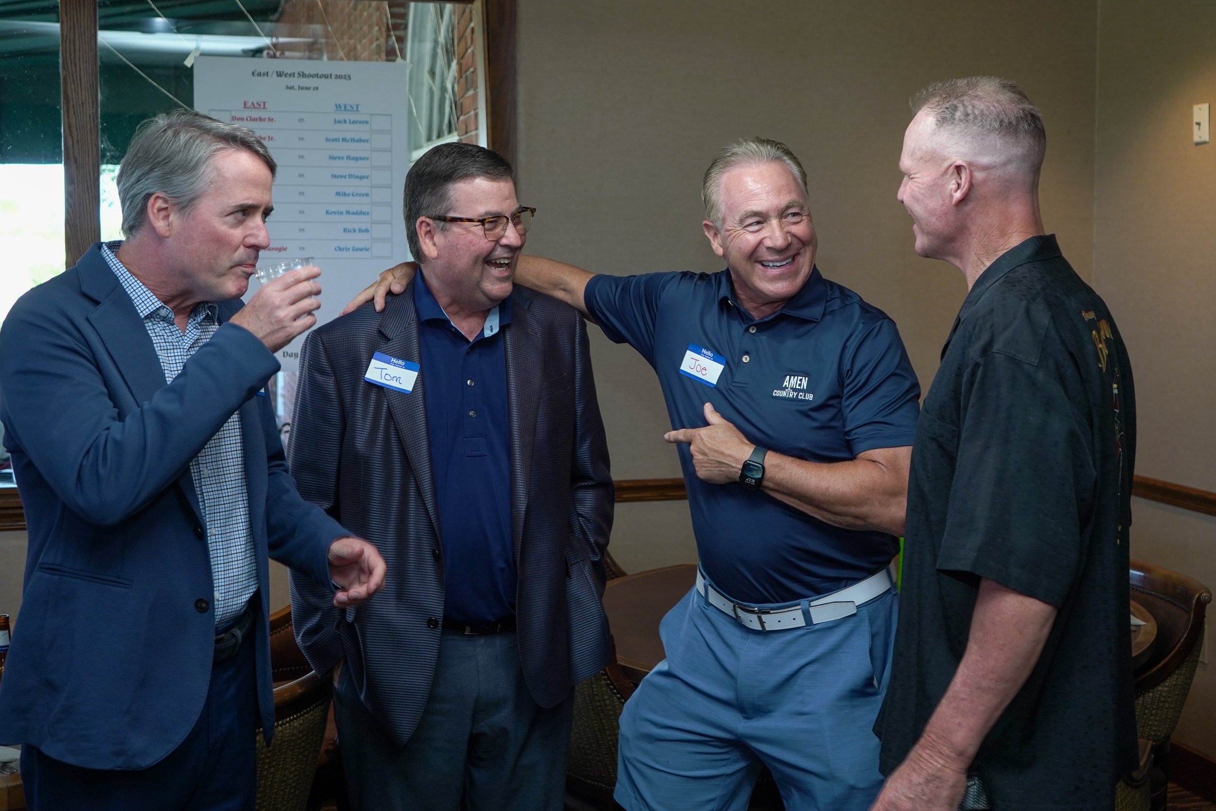 Four men, dressed in business casual and golf attire, are smiling and conversing in a room, with two of them wearing name tags that read 'Tom' and 'Joe.' They seem to be enjoying a friendly moment at a social or sporting event.