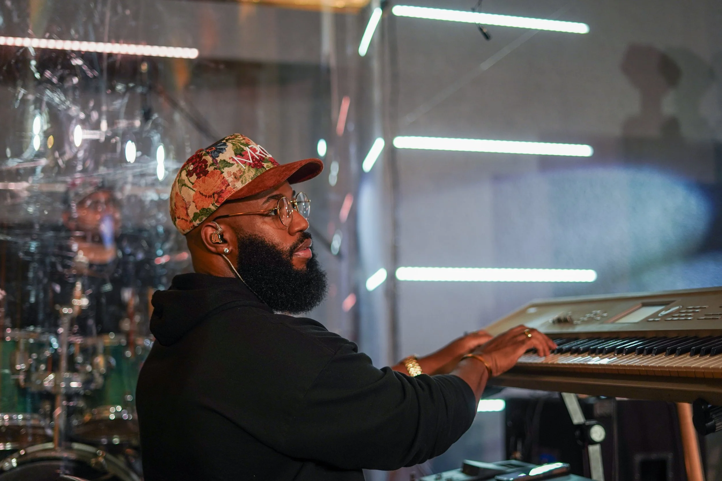 A man with glasses, a beard, and a floral cap is playing an electronic keyboard on stage, with a drummer visible in the background.