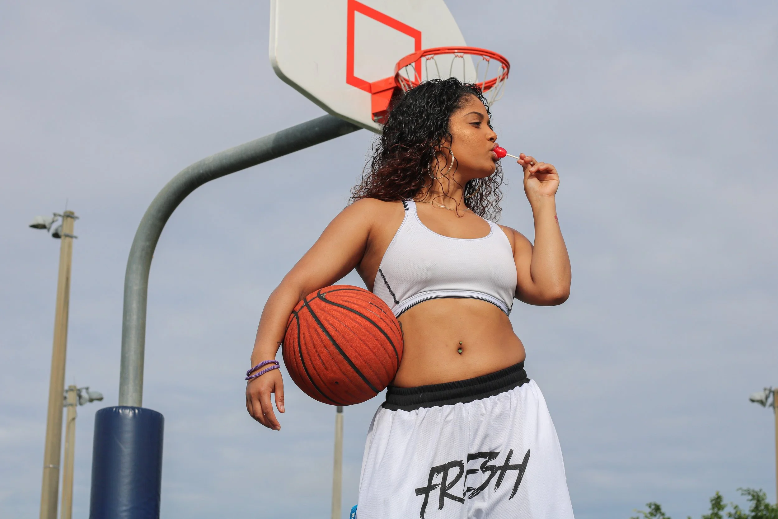 Young woman holding a basketball with one arm, standing on an outdoor basketball court, licking a red lollipop, wearing a white sports bra, white and black basketball shorts with the word 'FRESH' printed on them, and a belly button piercing.