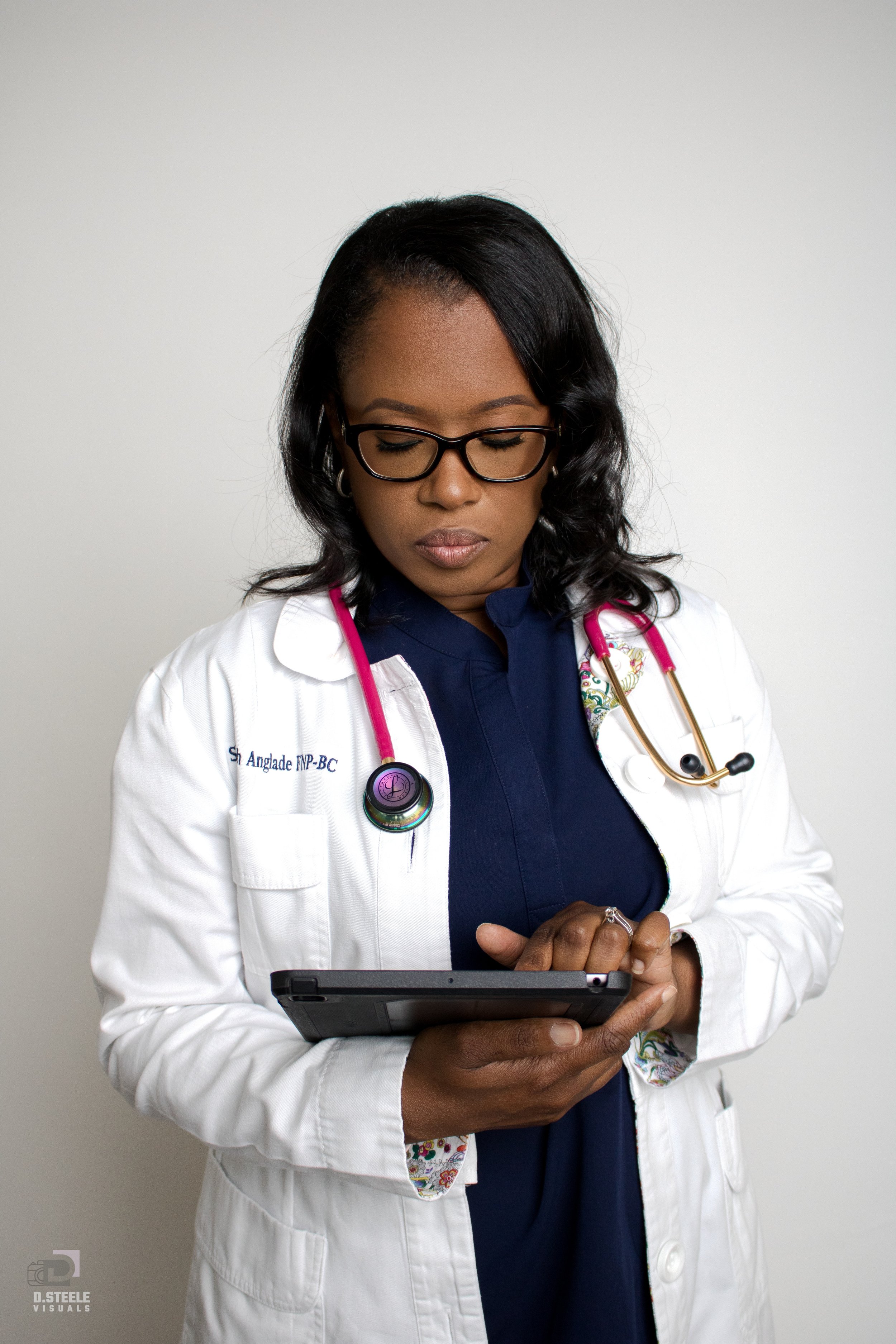 A female doctor with glasses wearing a white coat and a stethoscope, looking at a tablet device against a plain white background.