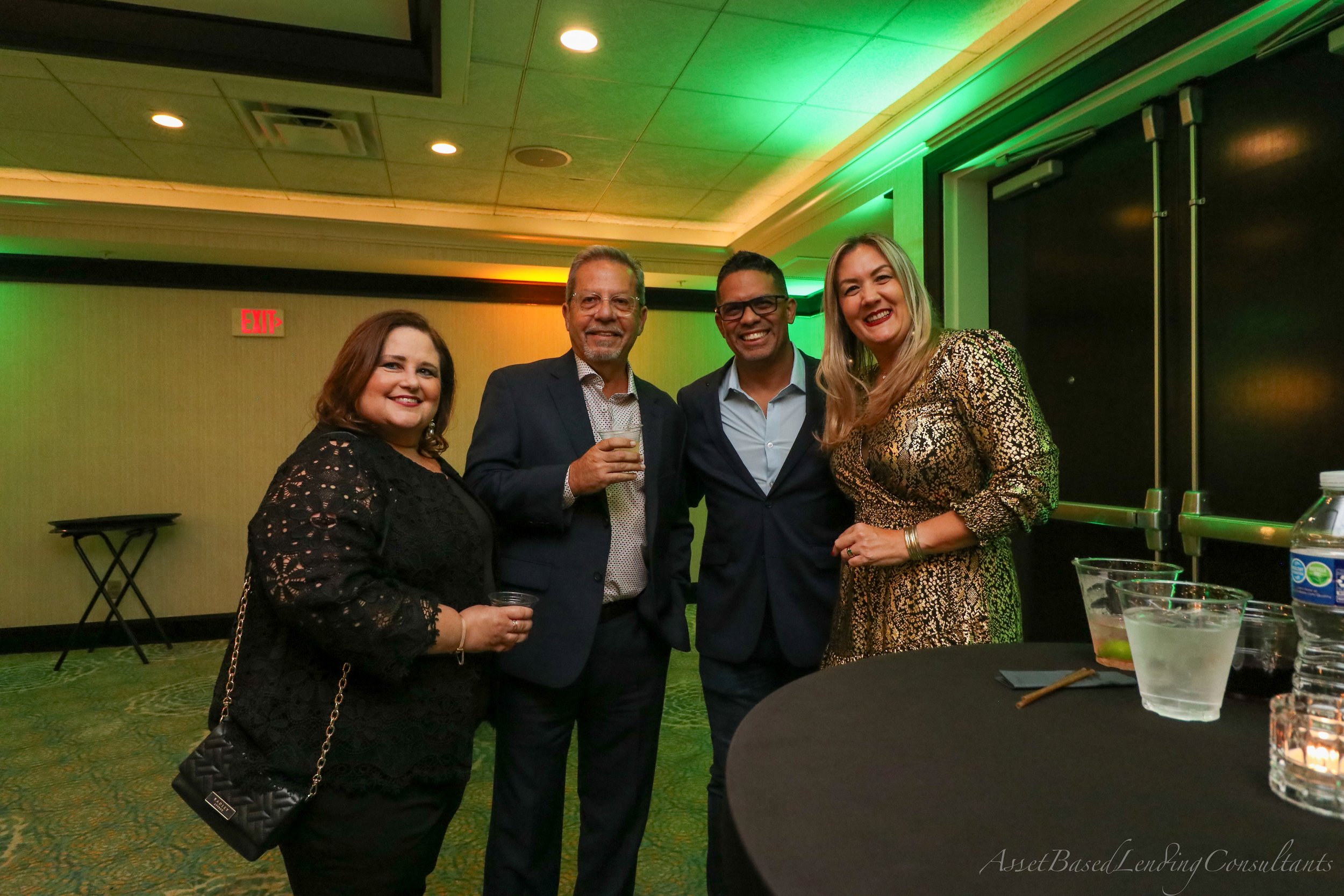 Four people smiling and posing together at a social event, standing inside a room with green ambient lighting, a black table with drinks, and a yellow wall.