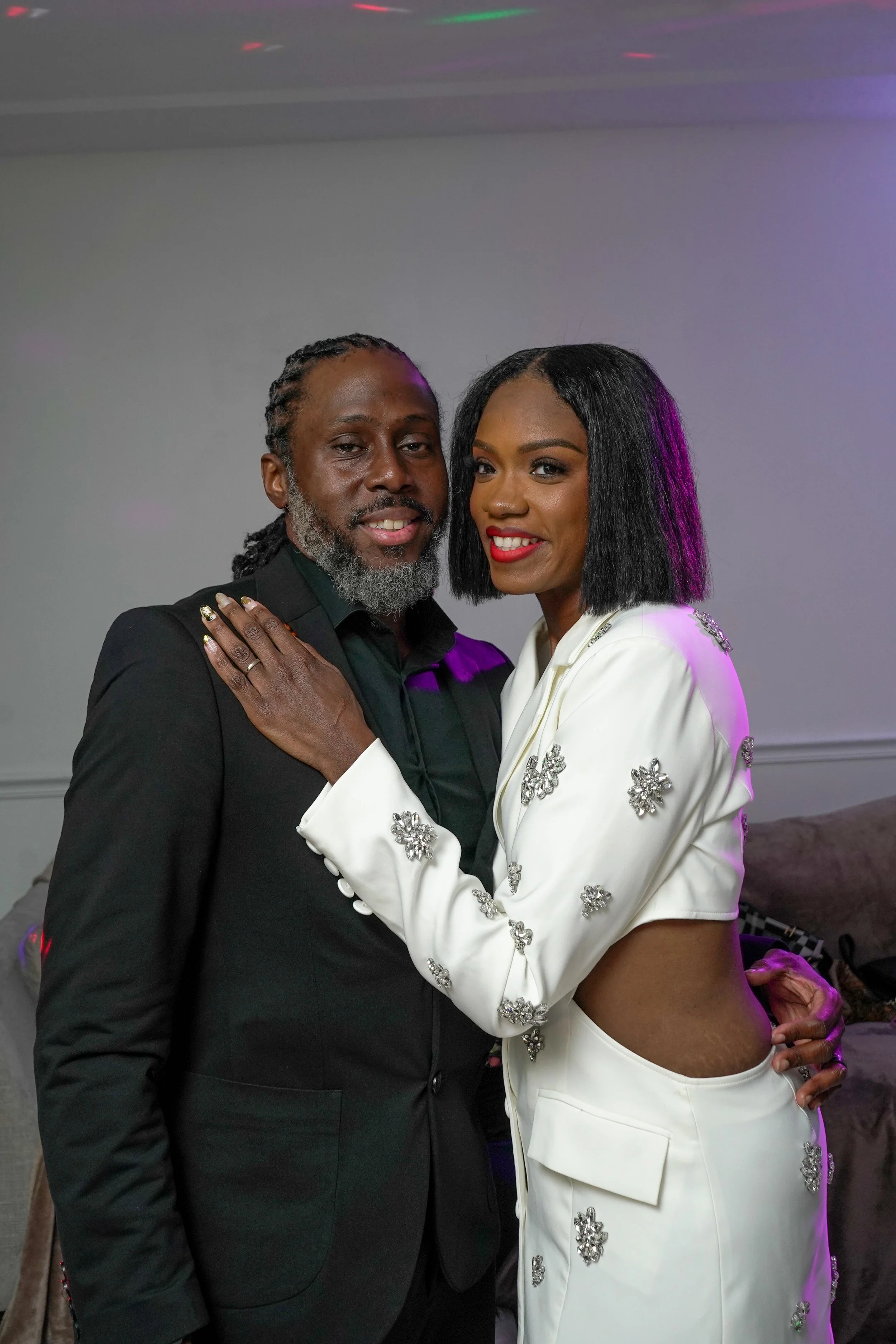 A man and woman pose together indoors. The man has gray hair and beard, and is wearing a black suit. The woman has black hair, red lipstick, and is dressed in a white suit with floral embellishments.