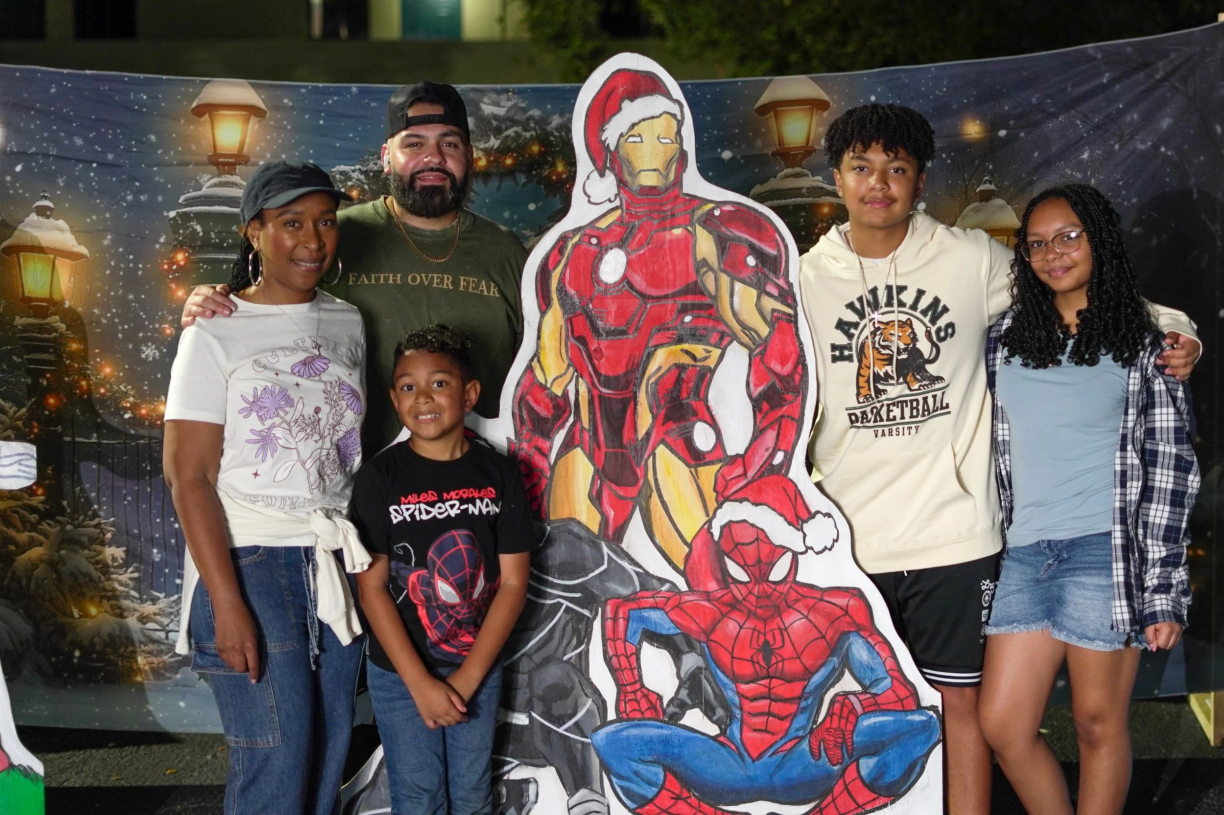A group of six people, including two adults and four children, standing together at a Christmas-themed event. They are posing with a large cutout of Iron Man and Spider-Man dressed in Christmas outfits, in front of a festive backdrop with snow, stree