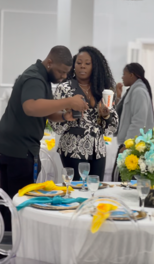 A woman holds a Dunkin' Donuts cup while looking at her phone, standing next to a man at a decorated event table with yellow and blue napkins, glassware, and a flower arrangement.
