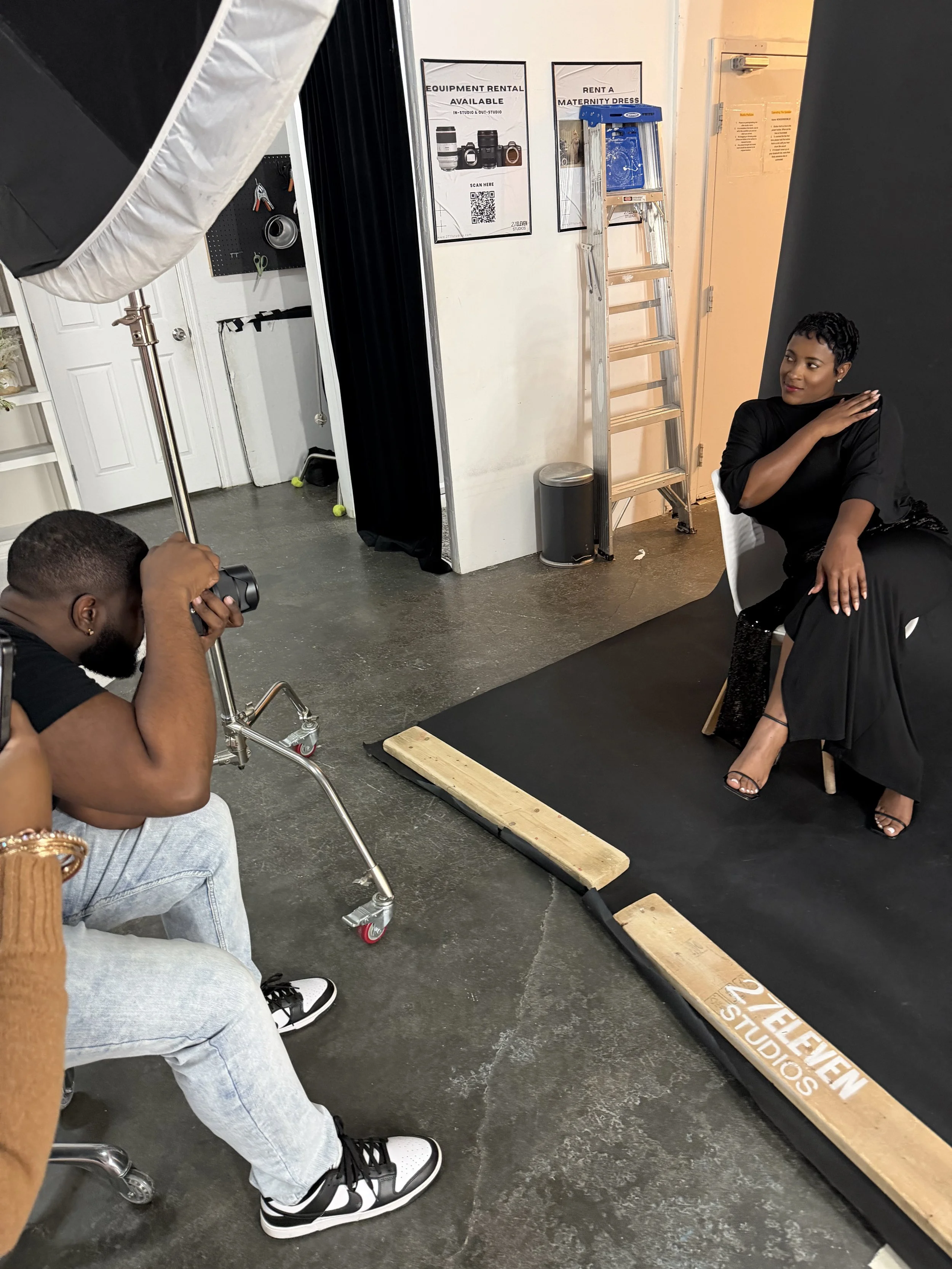 A photoshoot setup with a woman sitting on a chair, posing in front of a black backdrop, and a photographer capturing her image with a professional camera. There is studio lighting equipment and a ladder in the background.