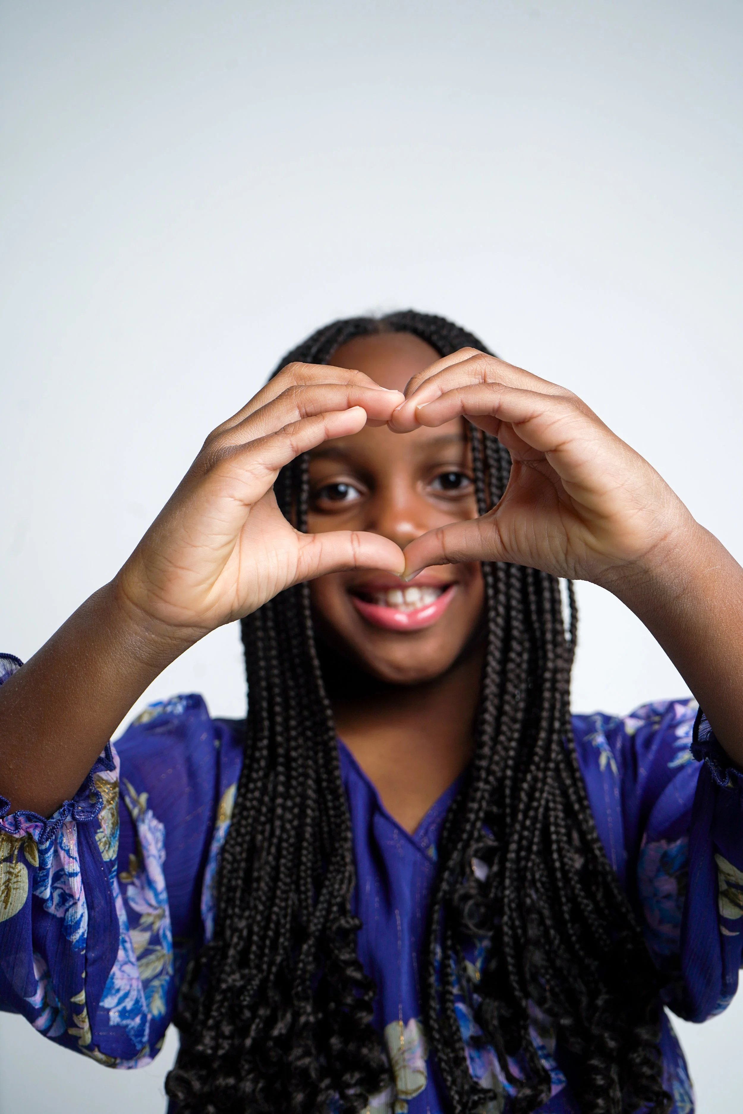 A young girl with braided hair making a heart shape with her hands and smiling.