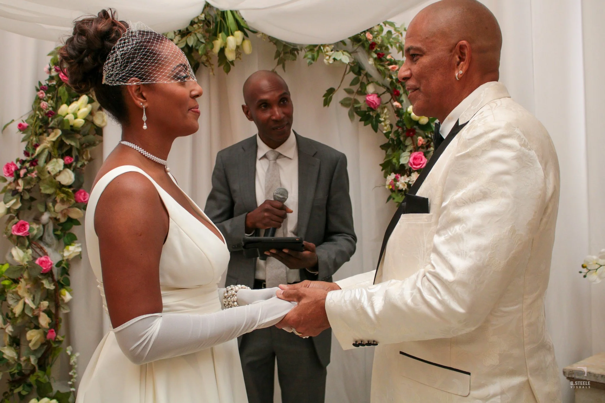 A bride and groom holding hands and looking at each other during their wedding ceremony, with a officiant standing behind them holding a microphone, in a decorated floral archway setting.