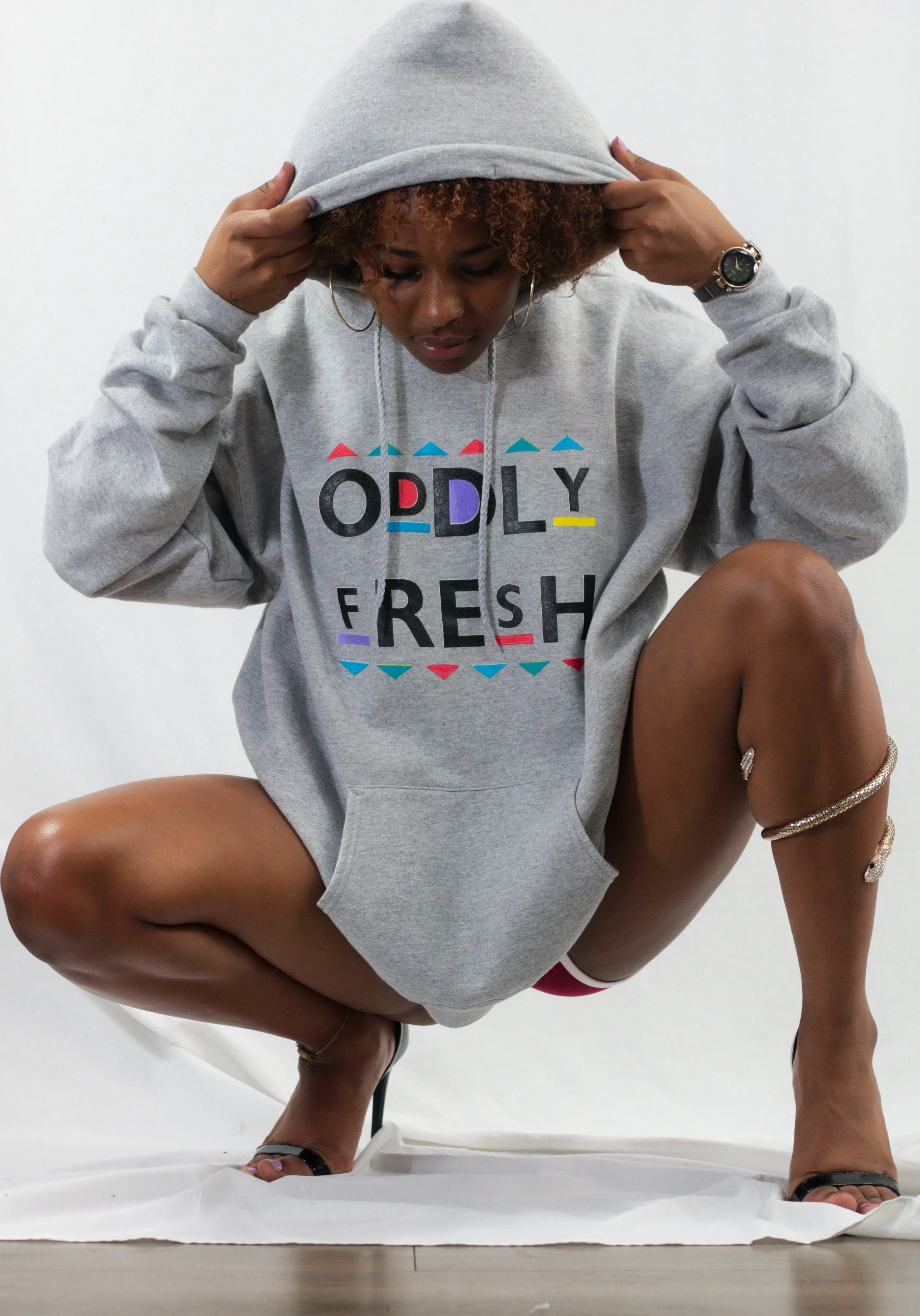 A woman in a grey hoodie with the words 'ODDLY FRESH' on it, squatting on the floor, looking down, with she has curly hair, some jewelry, and wearing high heels.