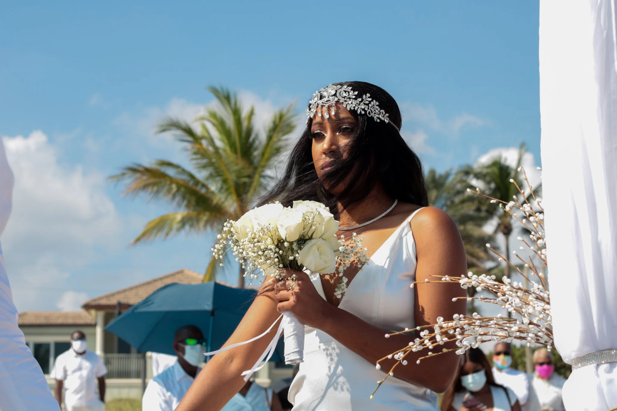 A woman with dark hair in a white dress holding a bouquet of white roses at an outdoor wedding ceremony, with palm trees and people wearing masks in the background.