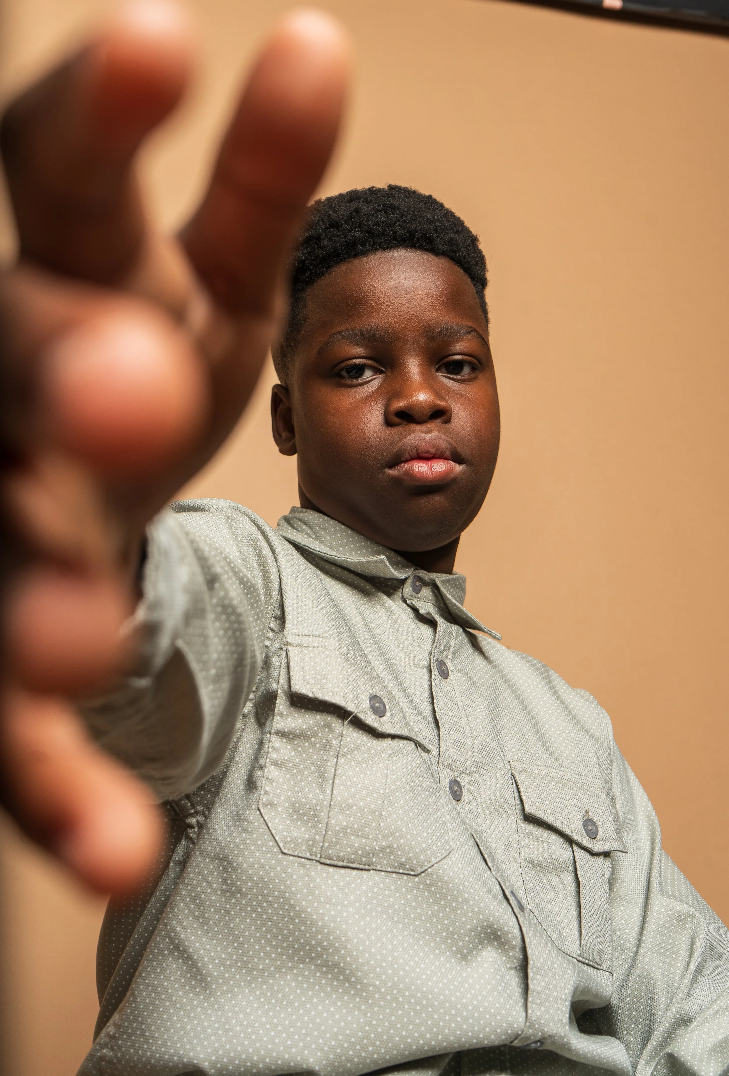 A young boy taking a selfie, wearing a light gray shirt with small dots and black buttons, with a neutral expression, against a beige background.