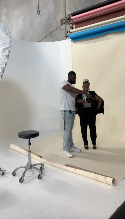 Man and woman standing on a beige backdrop in a photography studio, preparing for a photo shoot. The studio has a gray concrete wall and rolls of colored backdrop paper on the ceiling.