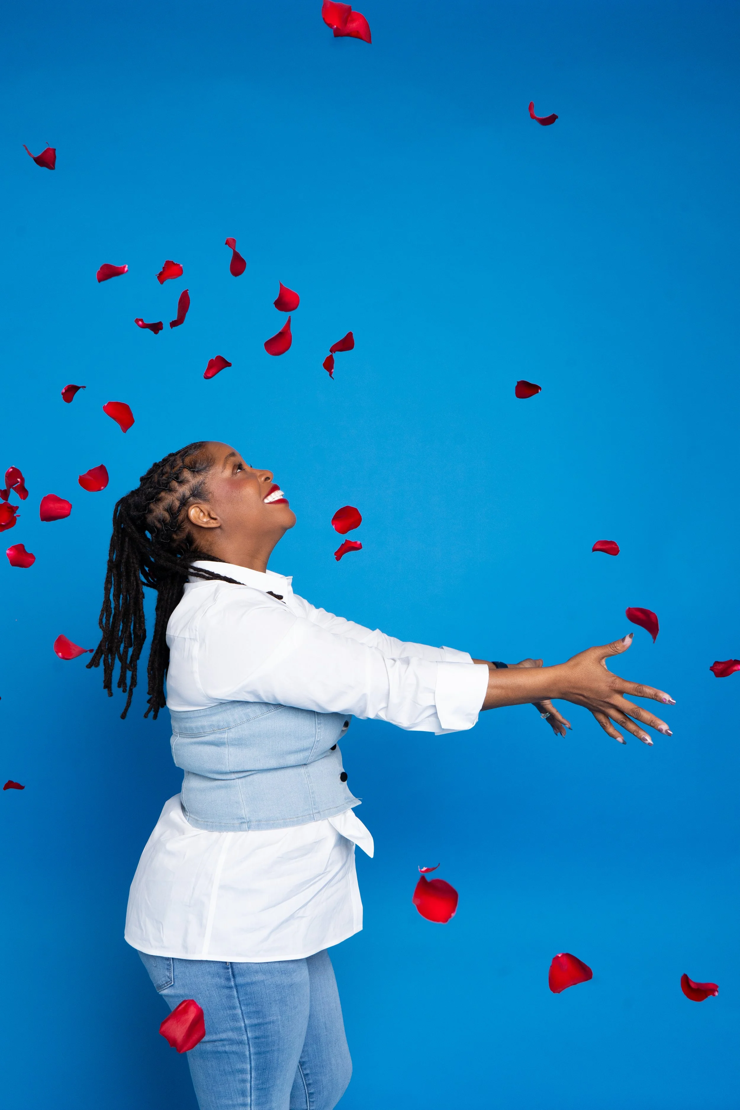 Woman with dreadlocks smiling with her arms outstretched as red rose petals fall around her against a bright blue background.