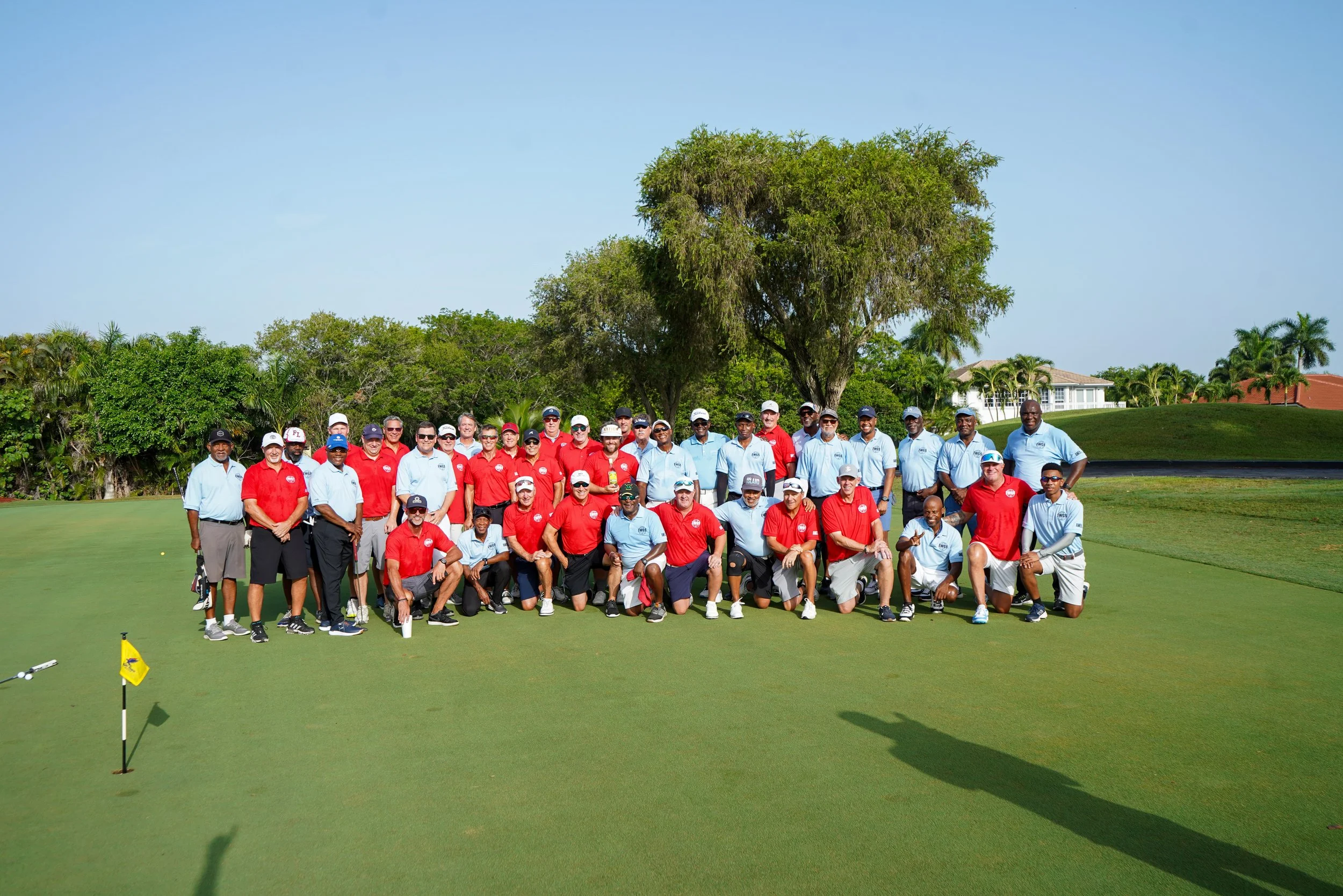 Group of people on a golf course, with green grass, trees, and houses in the background.