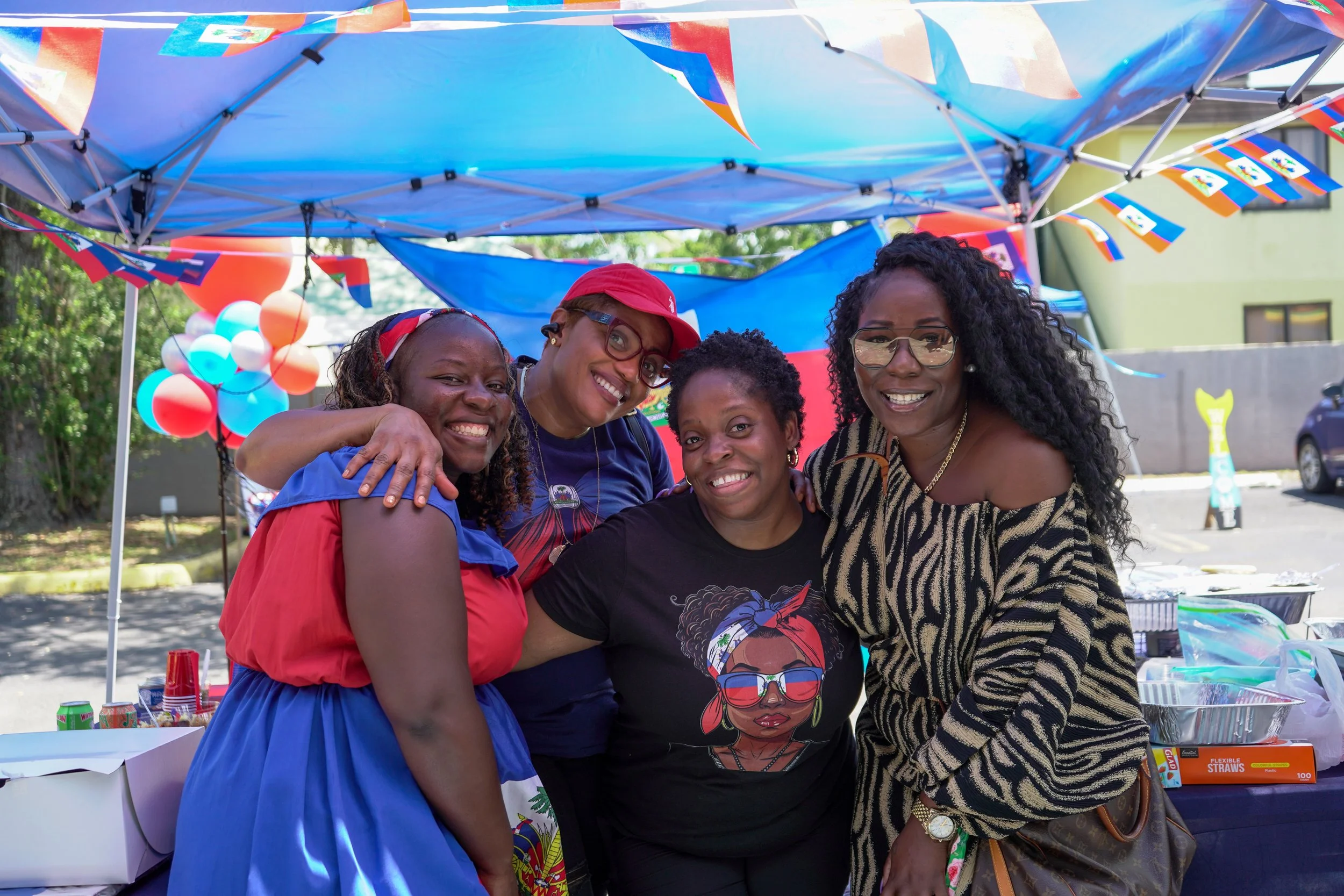 Four women smiling and hugging at an outdoor celebration under colorful banners and balloons, with a table of snacks and drinks nearby.