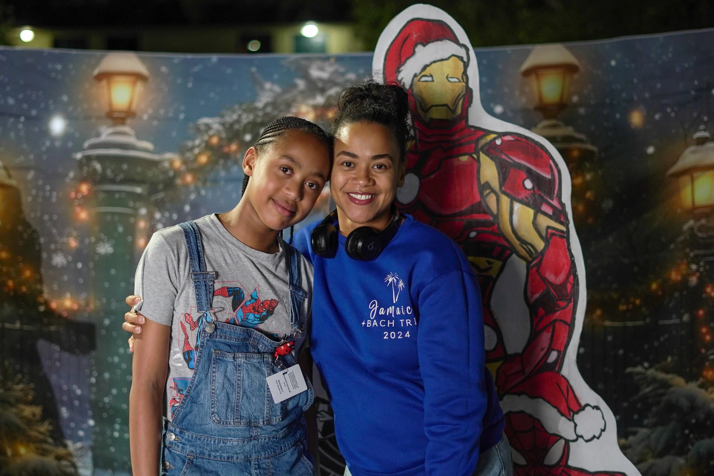 A woman and girl stand together smiling in front of a holiday-themed backdrop featuring Iron Man in a Santa hat, Christmas lights, and a snowy scene.