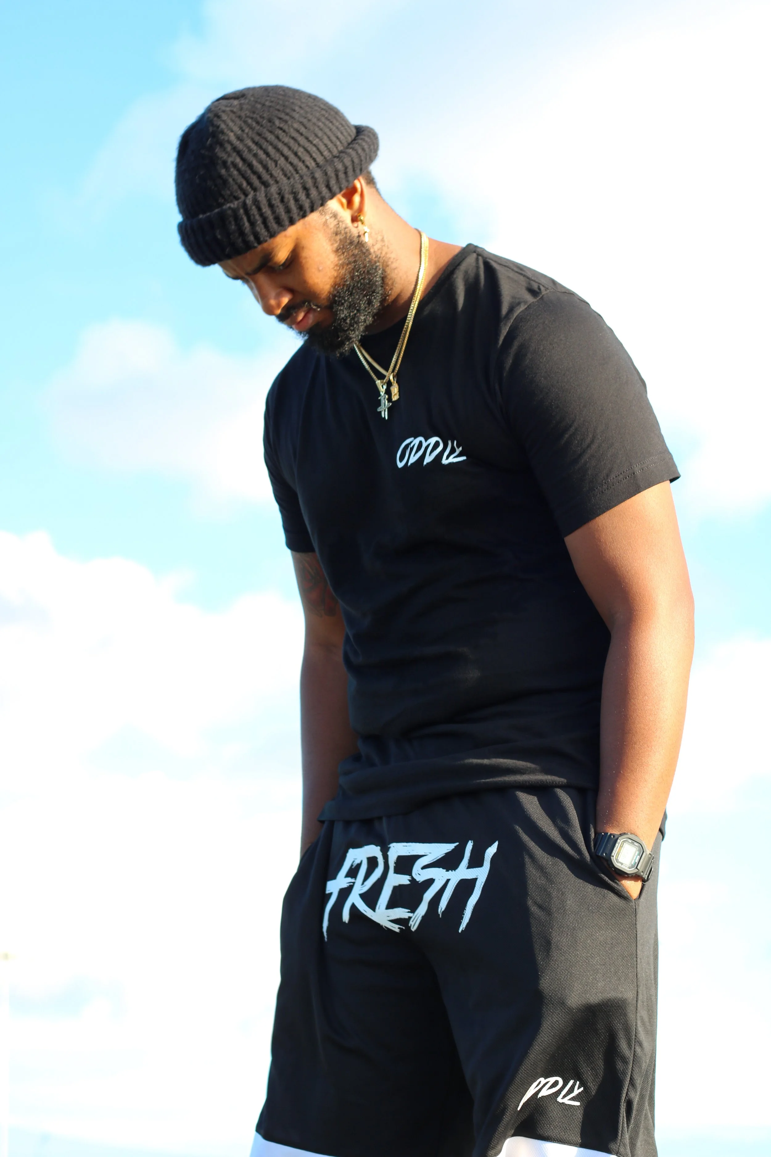 A man wearing a brown knit beanie, black t-shirt, and black shorts with the words 'FRESH' and 'ODDL' on them, looking down against a backdrop of blue sky with clouds.