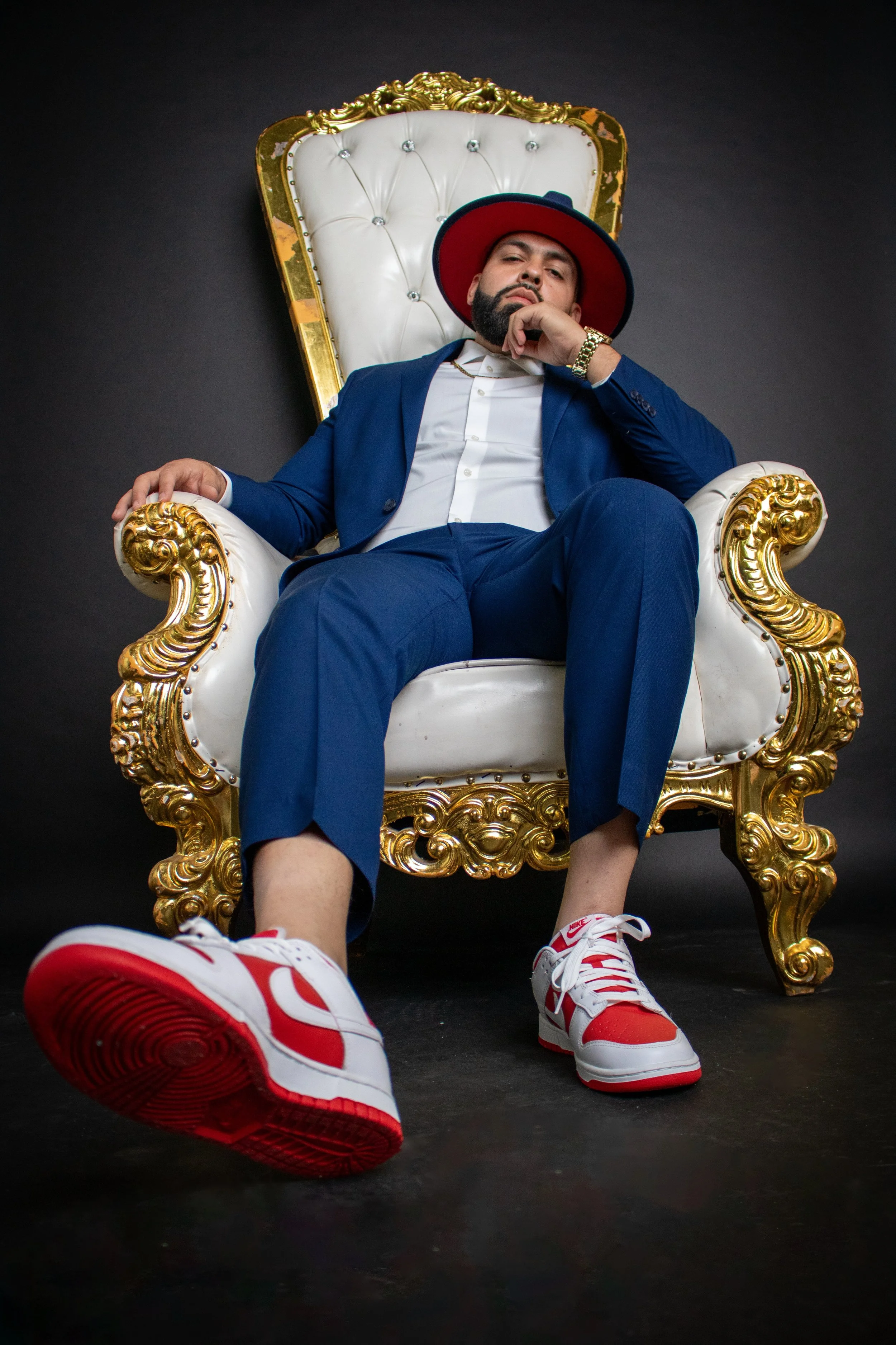 A man in a blue suit, white shirt, red and white Nike sneakers, wearing a red and blue hat, sitting on an ornate white and gold throne against a dark background.
